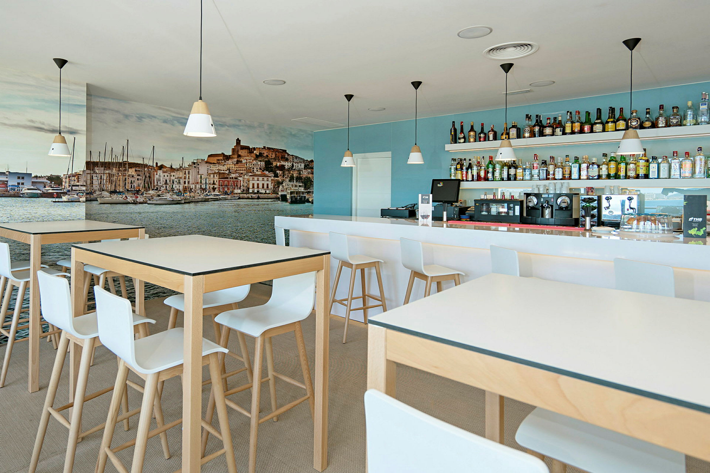 En moderne kafé med høye bord og stoler, en bar med drikkevarer i bakgrunnen og et stort veggmaleri av en havneby. (AI-generert tekst)