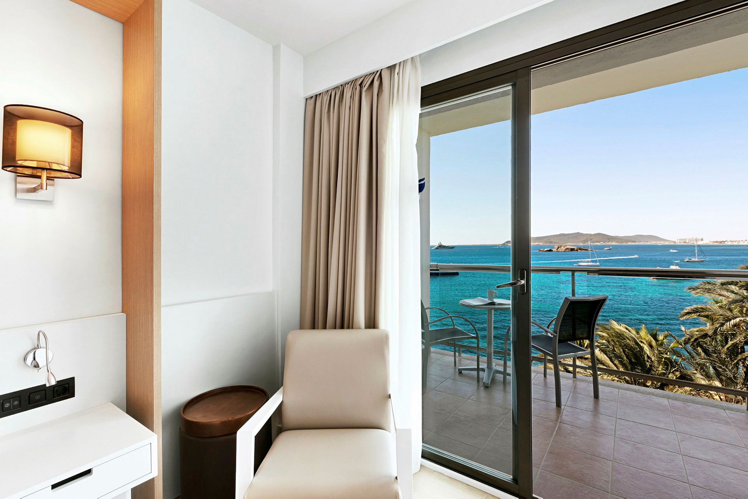 Hotellrom med minimalistisk innredning og utsikt over havet fra en balkong. (AI-generert tekst)