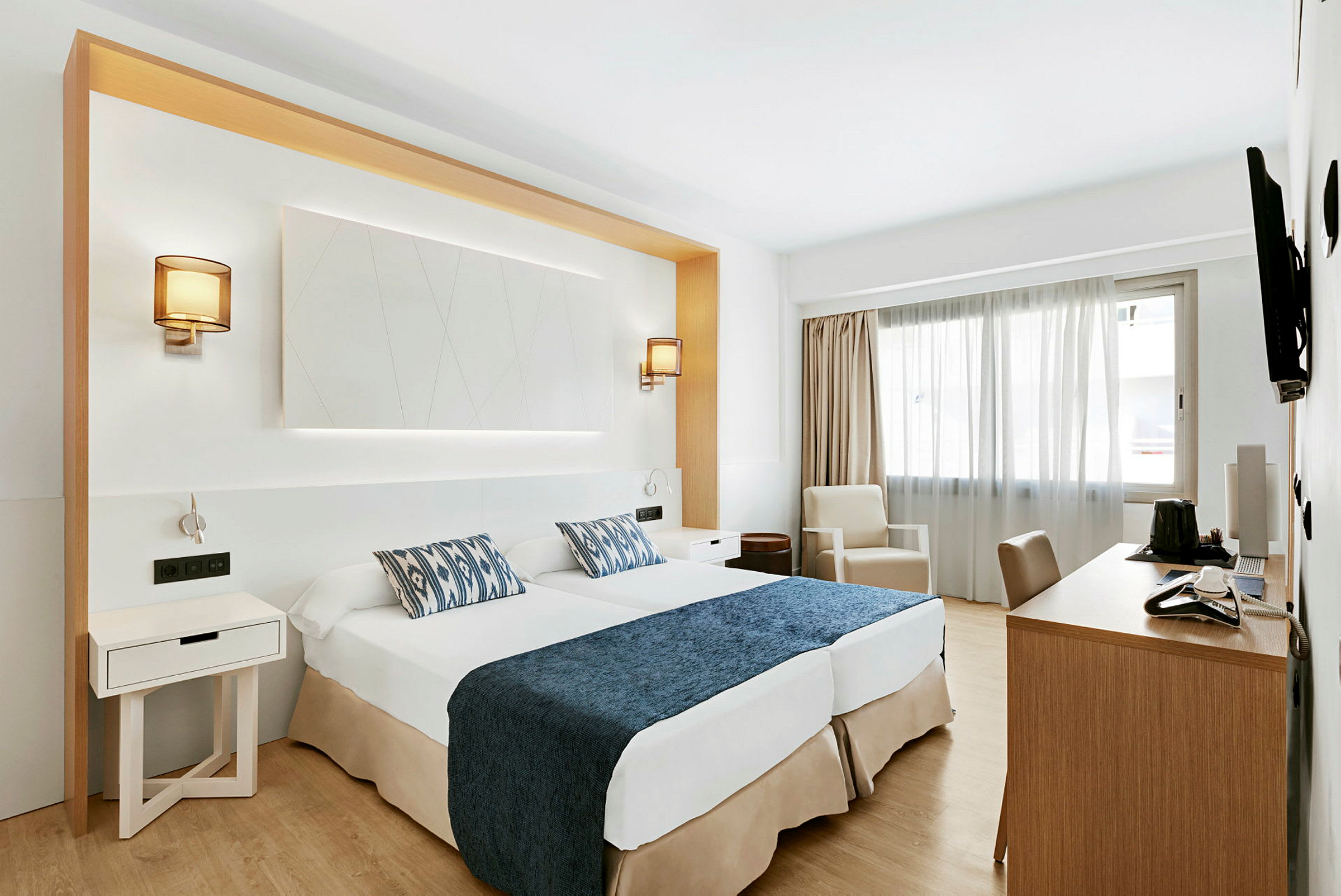 Et hotellrom med to senger med blått sengeteppe og mønstrede puter, skrivebord, stol og en lenestol ved et vindu. (AI-generert tekst)