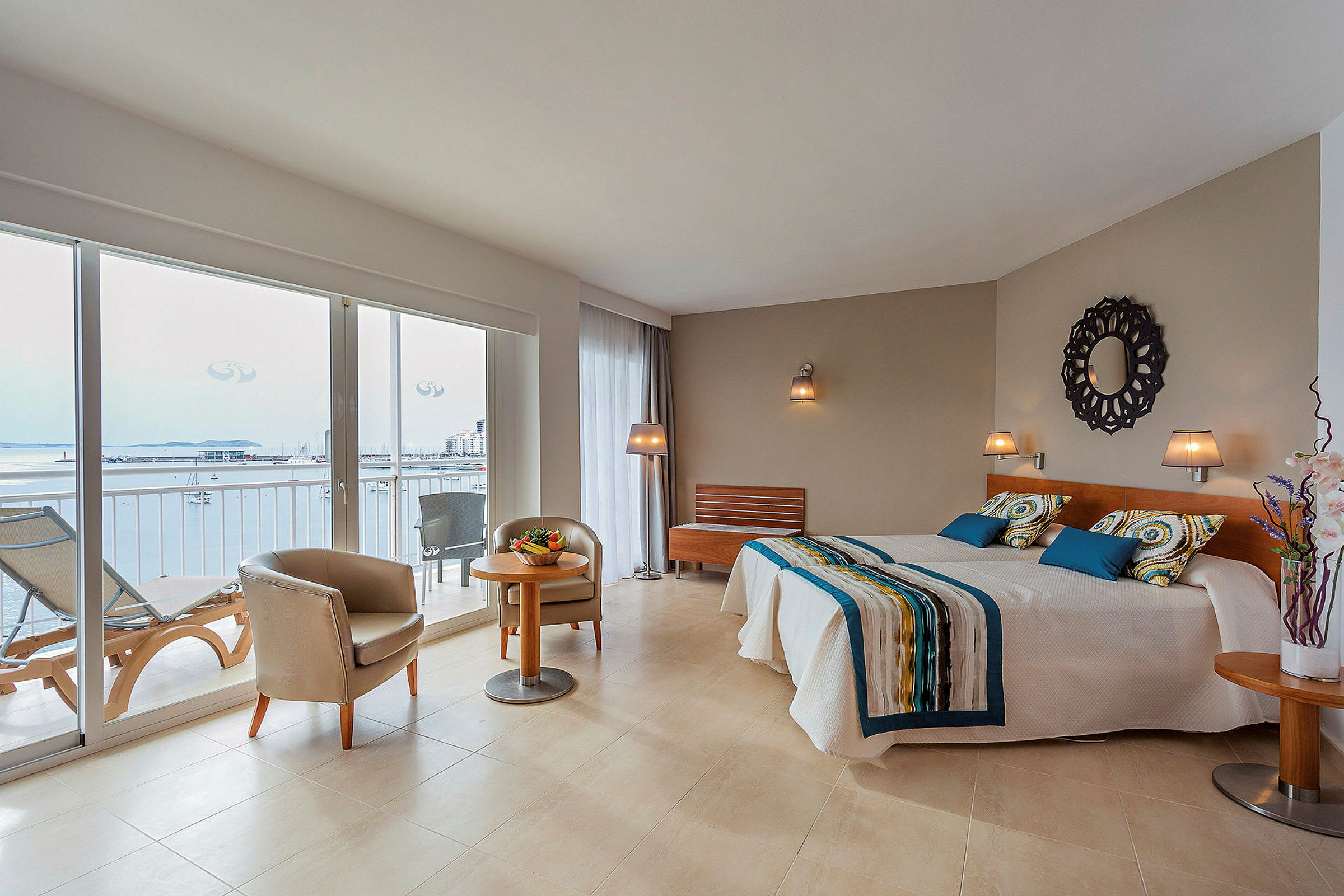 Et hotellrom med store glassvinduer som viser en balkong og utsikt mot havet. Det er to enkeltsenger, en sittegruppe og moderne innredning. (AI-generert tekst)