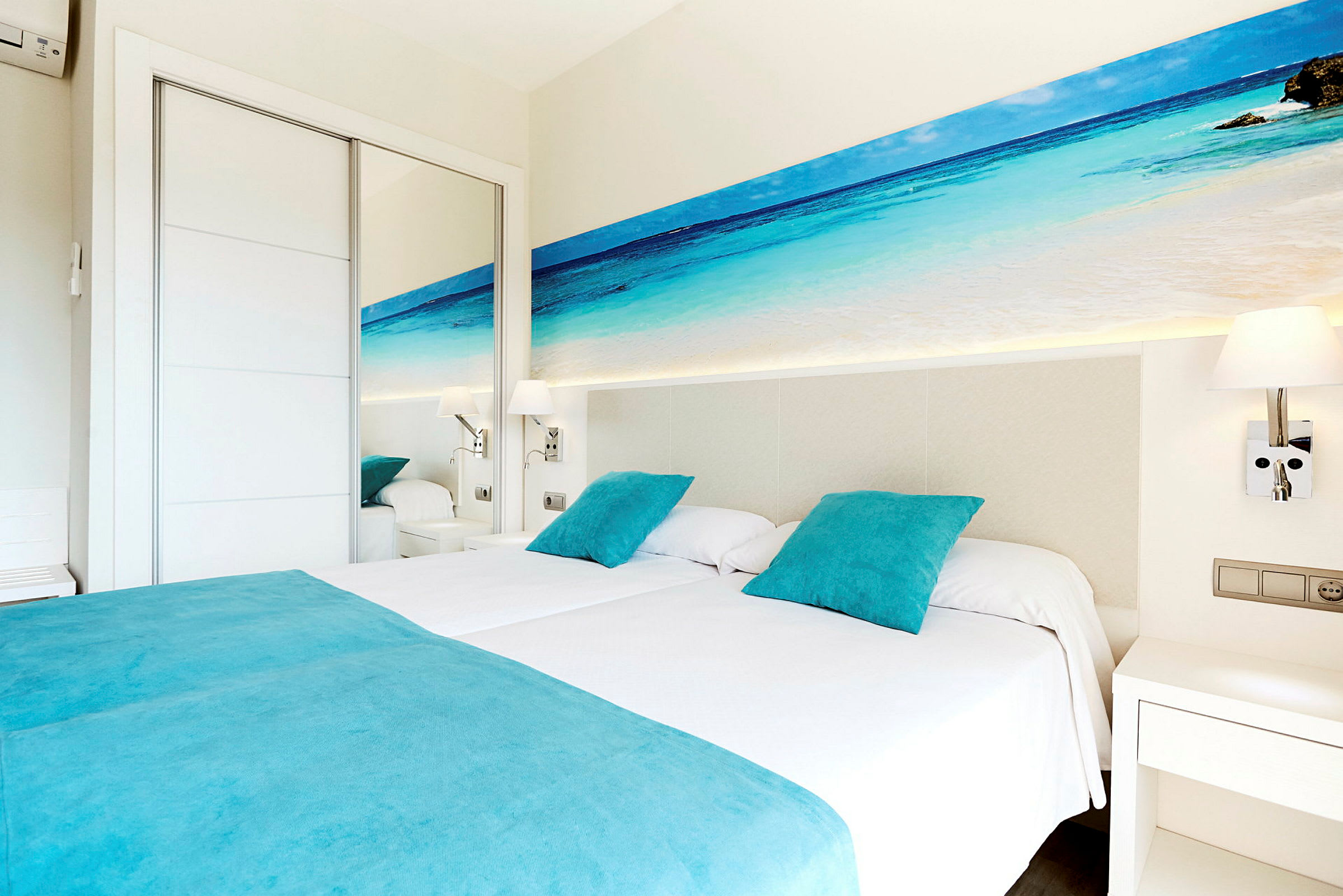 Et hotellrom med to enkeltsenger, oppredde med hvitt sengetøy og turkise puter og sengeteppet. På veggen over sengene er det et bilde av en strand og havet. Rommet har hvite møbler og et speil. (AI-generert tekst)