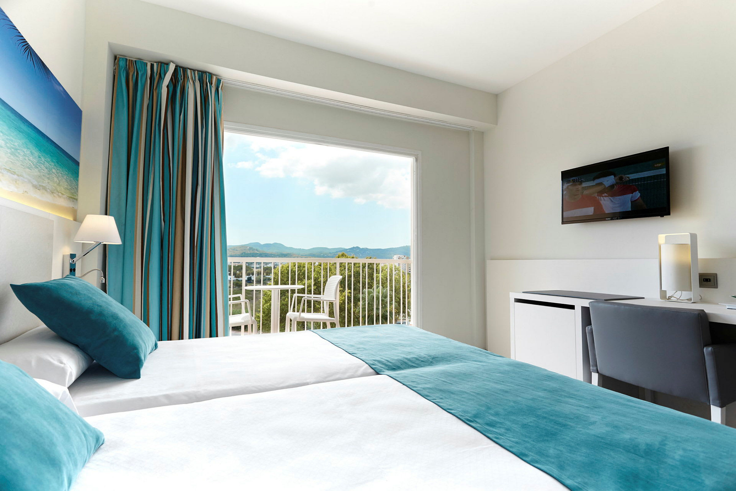 Et hotellrom med to enkeltsenger, et maleri med strandmotiv på veggen, store vinduer med utsikt over landskapet, og en TV på veggen. (AI-generert tekst)