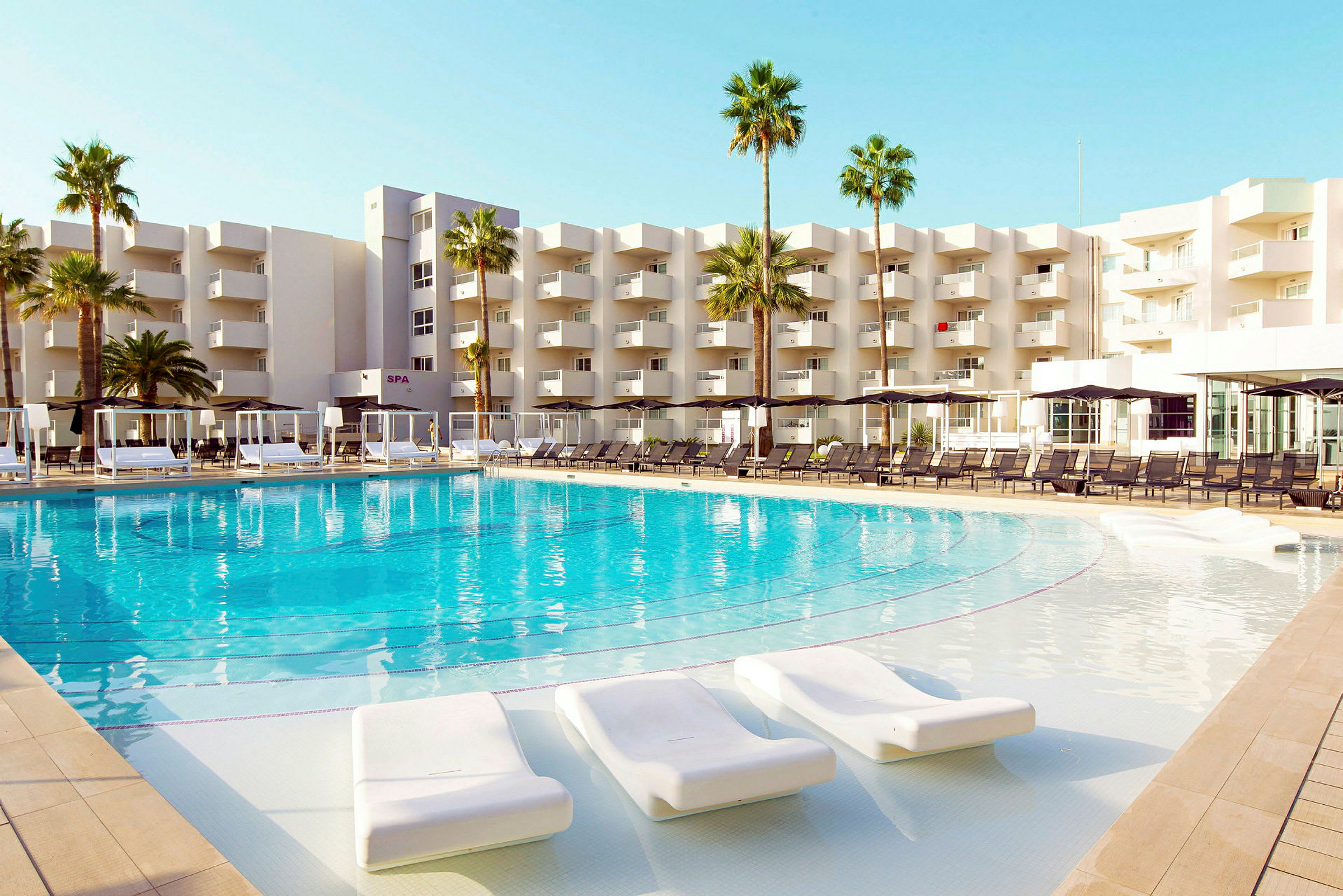 Stor svømmebasseng med solsenger rundt, foran en moderne hotellbygning og palmer. (AI-generert tekst)