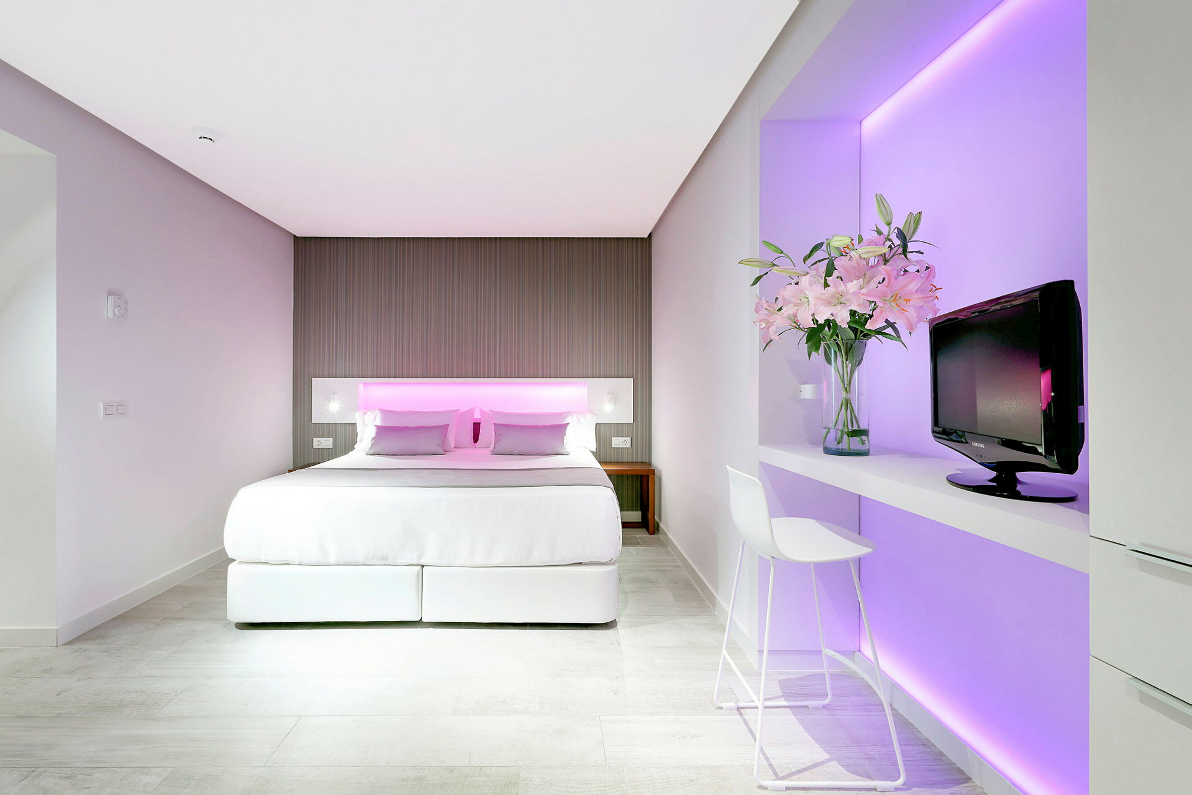 Hotellrom med dobbeltseng, elegant interiør og rosa belysning bak sengegavlen. En TV og en vase med blomster står på en hylle ved siden av. (AI-generert tekst)