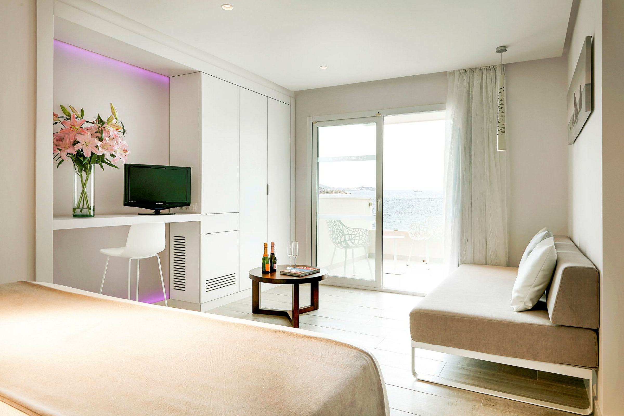 Et lyst hotellrom med en seng, en sofa, et bord og en balkong vendt mot havet. (AI-generert tekst)