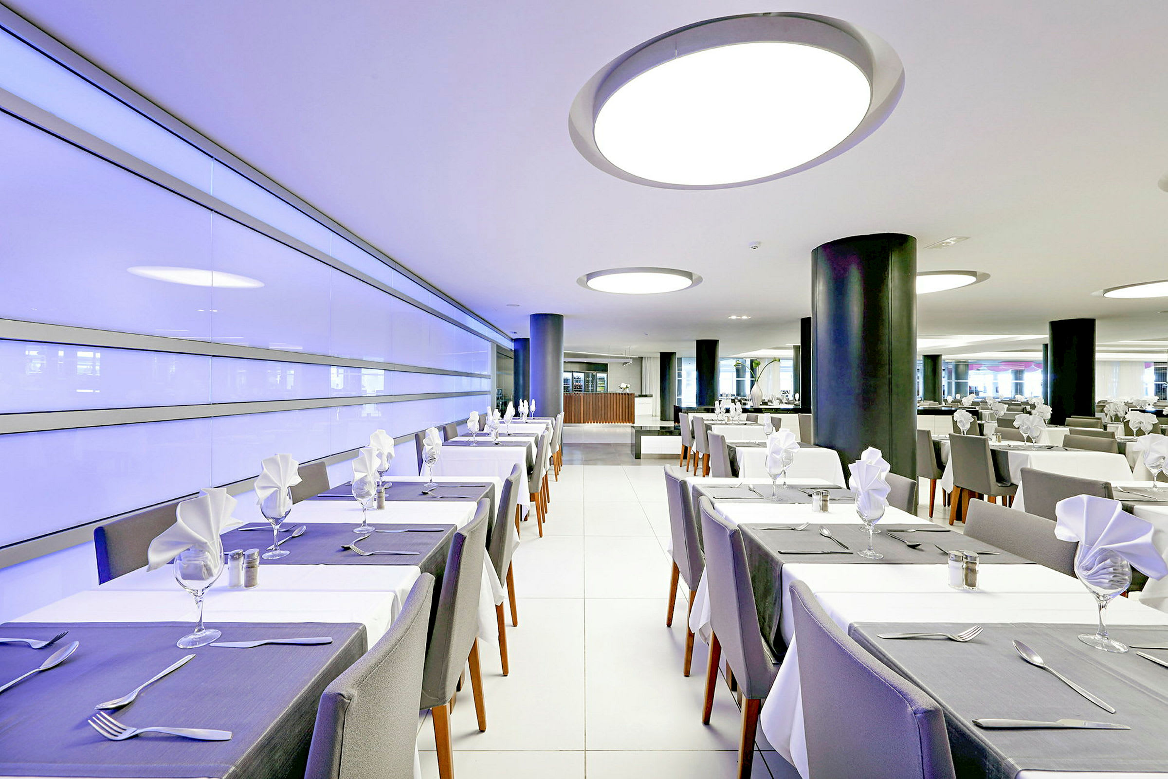 En moderne restaurant med dekkede bord og hvite stoler. Interiøret inkluderer moderne lamper og en lys atmosfære. (AI-generert tekst)