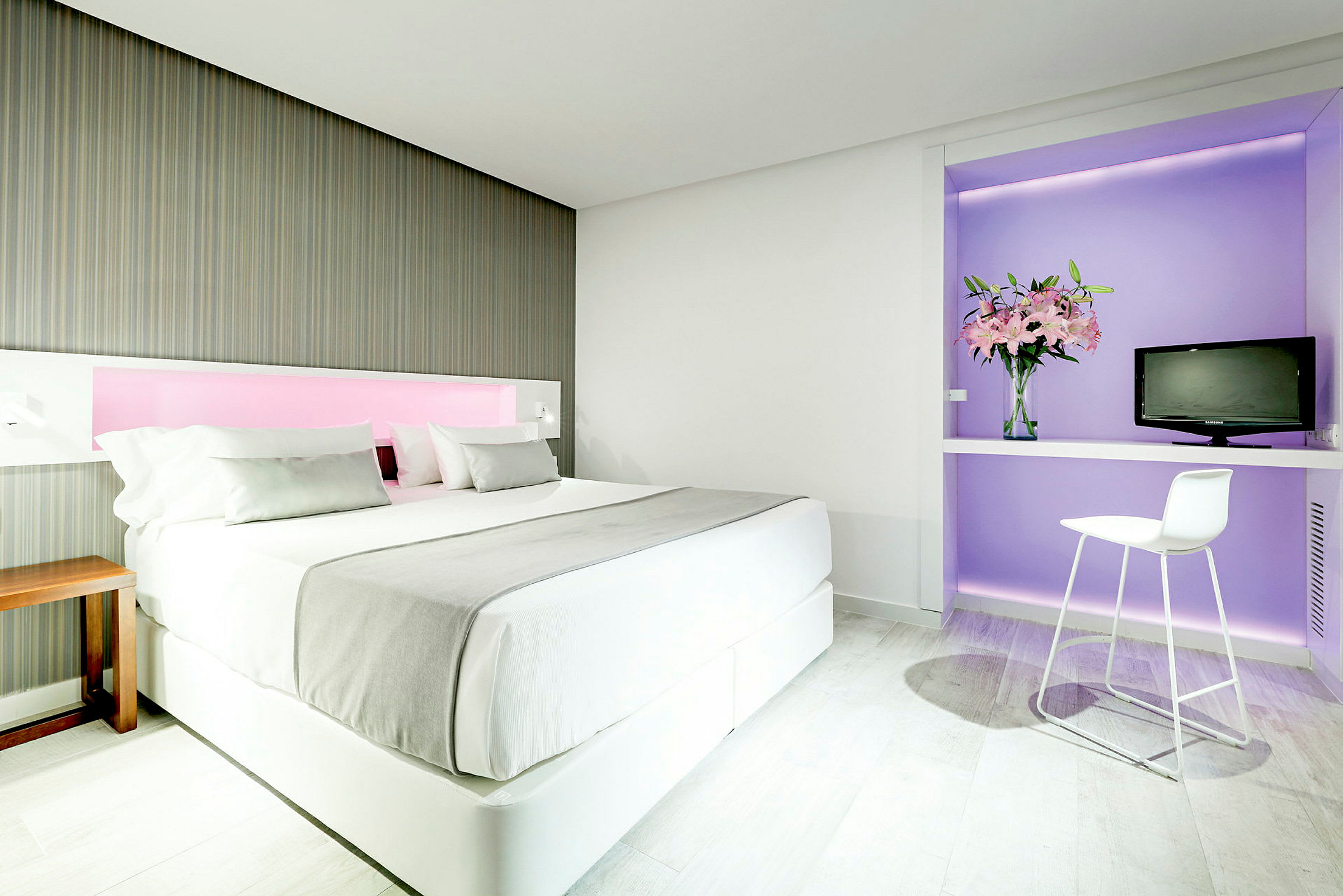 Et moderne hotellrom med en stor seng, dekorert i lyse farger. Til høyre er et skrivebord med en stol, en vase med blomster og en liten TV. (AI-generert tekst)
