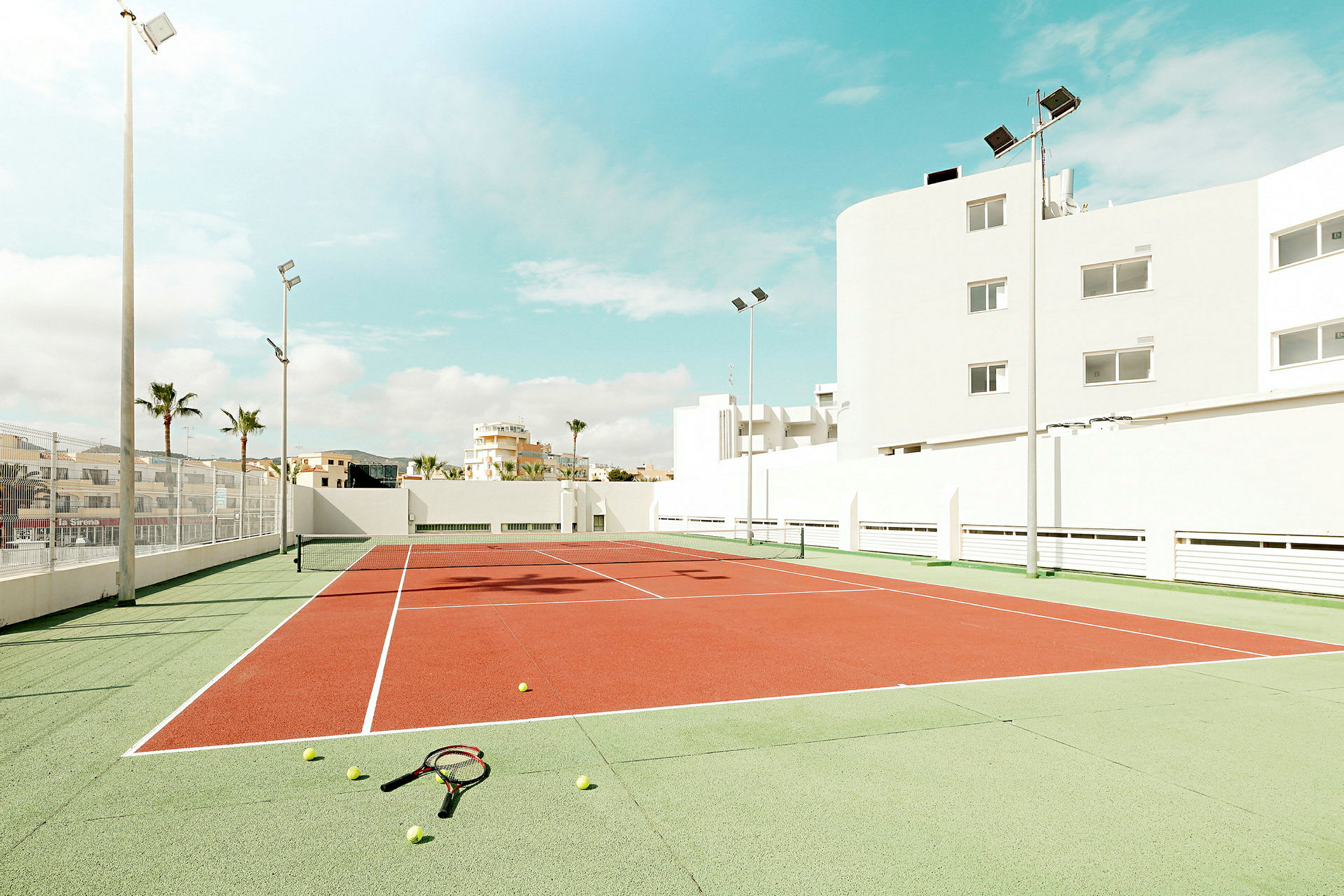En tennisbane omgitt av bygninger under en solrik himmel, med tennisballer og racketer liggende på bakken. (AI-generert tekst)