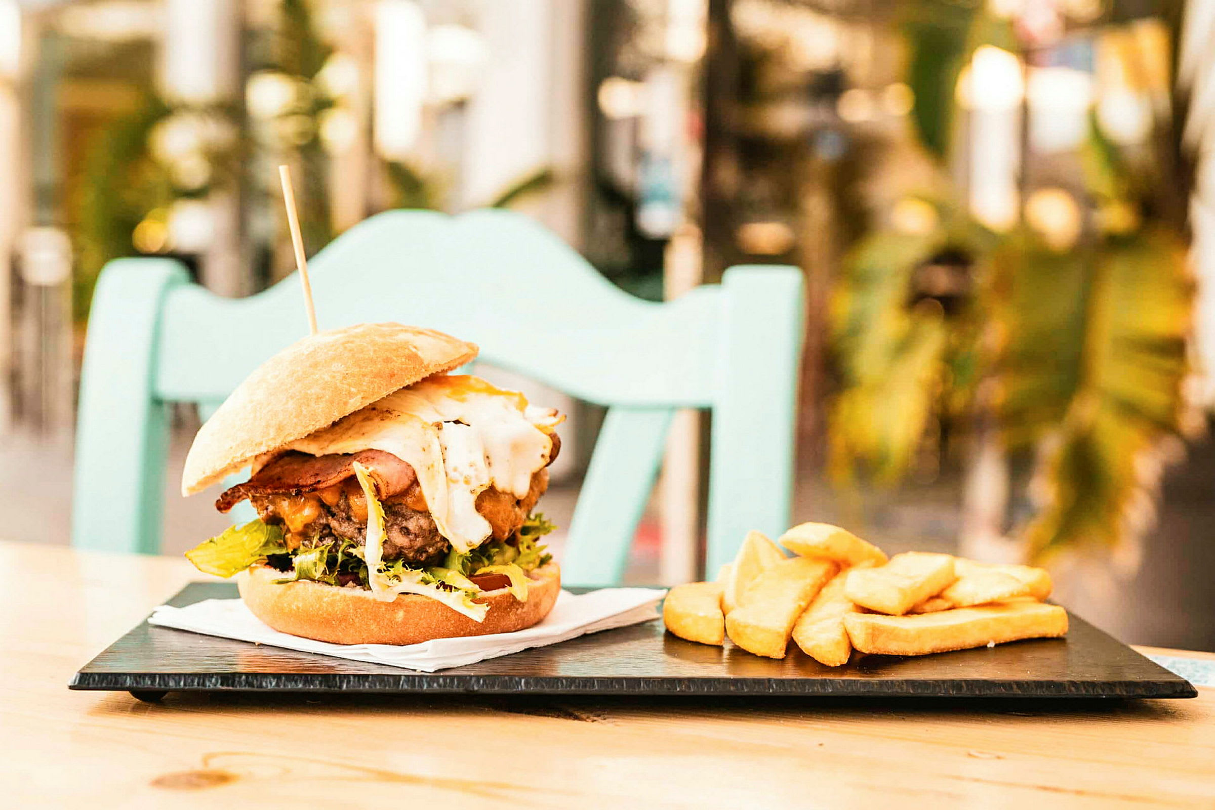En hamburger med tilbehør av pommes frites på en tallerken, plassert på et bord med en stol i bakgrunnen. (AI-generert tekst)