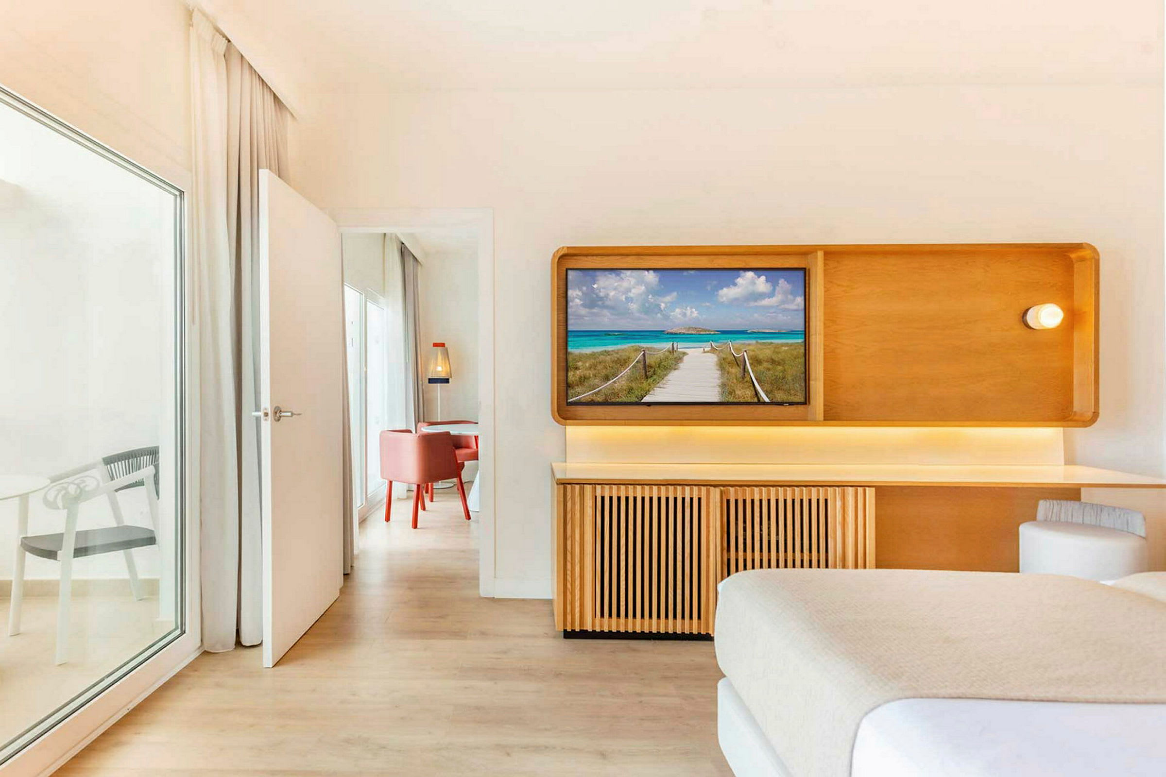 Et hotellrom med en TV som viser en strandpromenade. En dør fører til et tilstøtende rom med røde stoler. (AI-generert tekst)