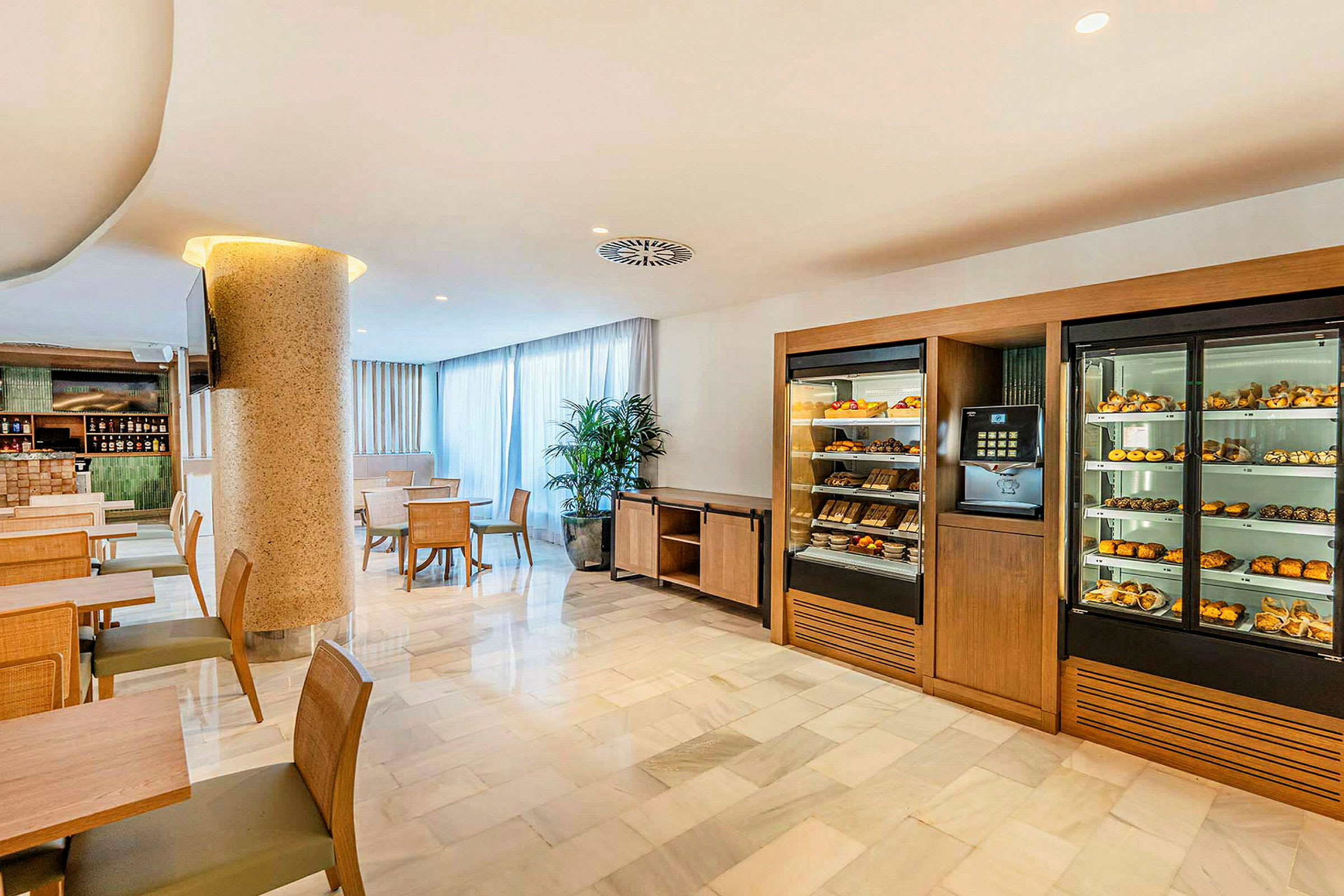 En hotellrestaurant med bord og stoler samt skap fylt med bakverk og frukt. (AI-generert tekst)