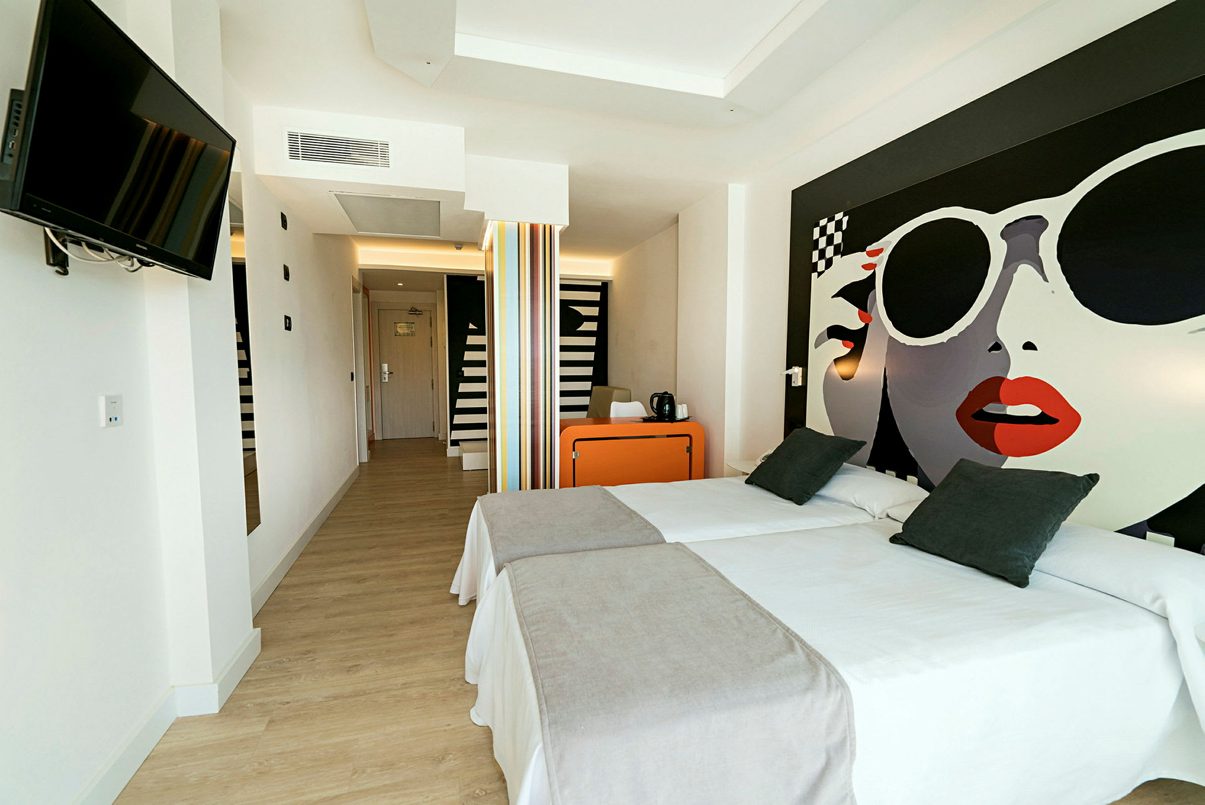 Hotellrom med to enkeltsenger, et oransje skrivebord og en veggmontert TV. (AI-generert tekst)