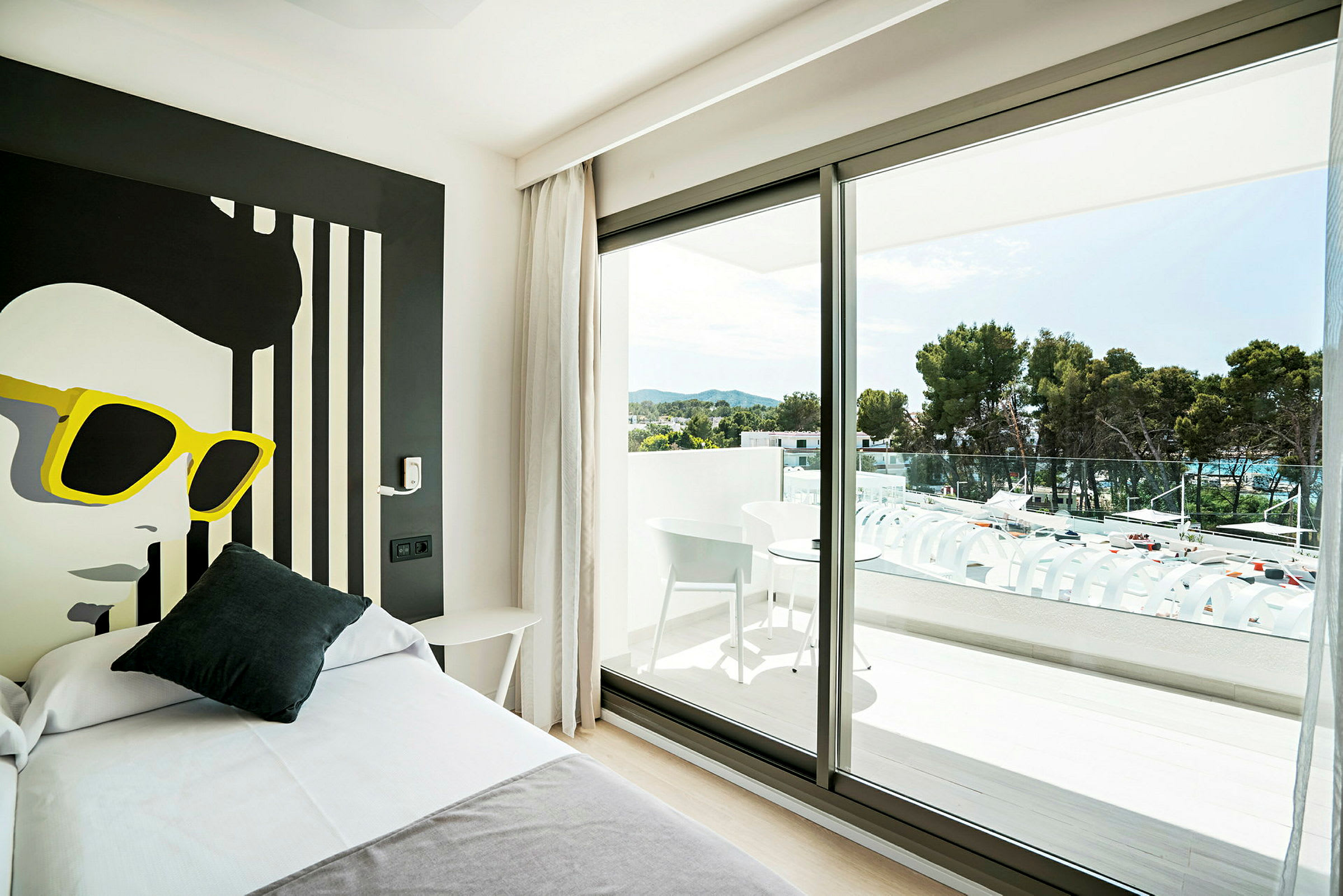 Et hotellrom med moderne innredning, en seng i forgrunnen og et stort vindusparti som leder ut til en balkong med møbler og utsikt over grønne trær. (AI-generert tekst)