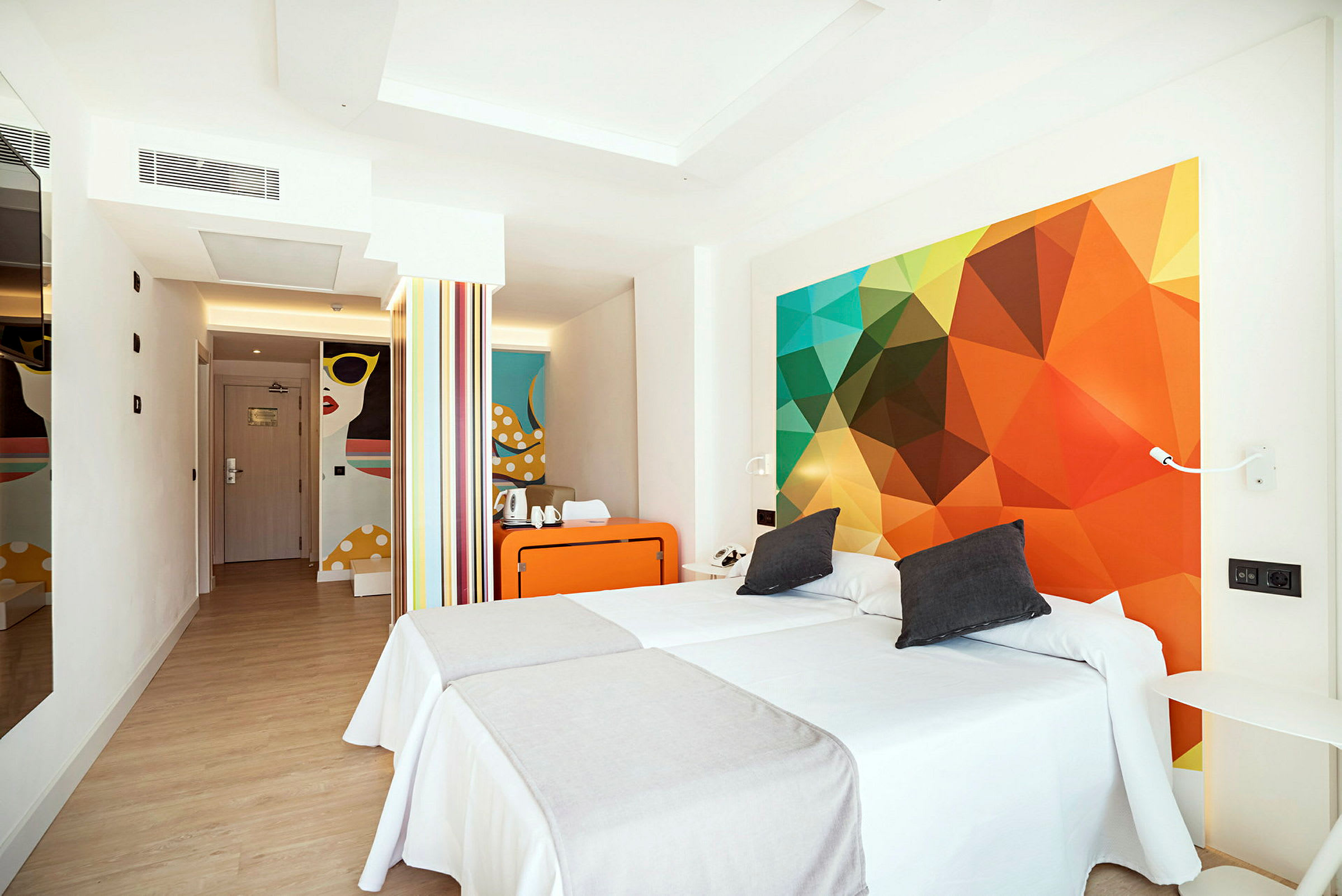 Et moderne hotellrom med to enkeltsenger, fargerike veggdekorasjoner og en liten sitteplass. (AI-generert tekst)