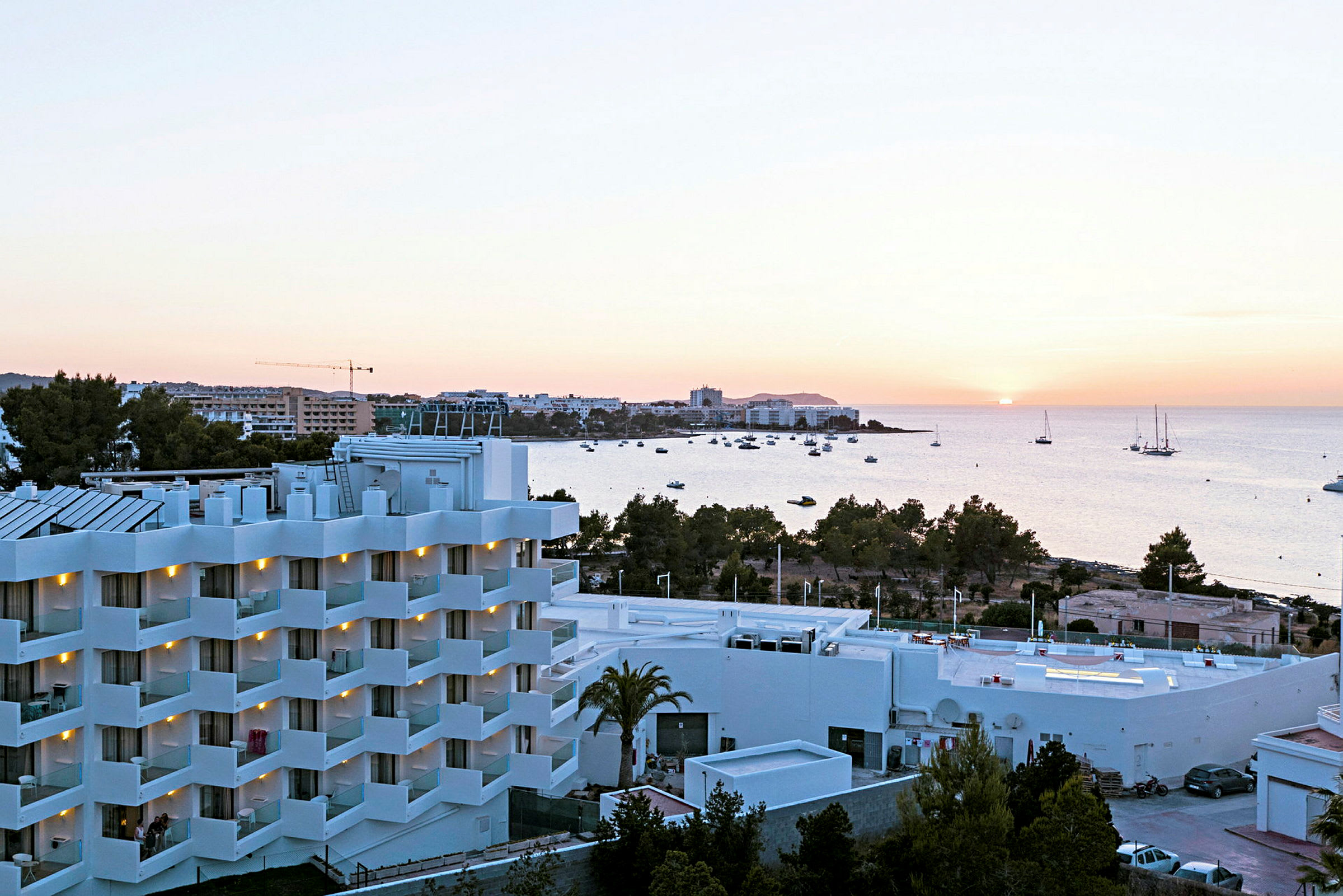 En utsikt over et hvitt hotell med balkonger, omgitt av grøntområder, med havet og en solnedgang i bakgrunnen. (AI-generert tekst)