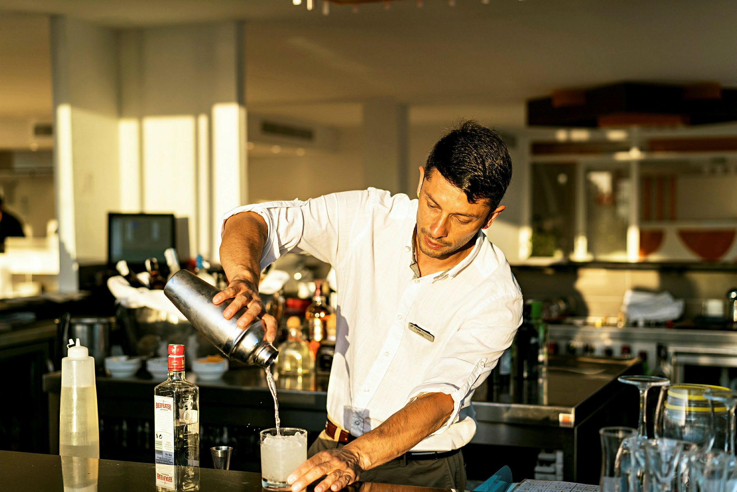 En person heller væske fra en cocktailshaker i et glass. I bakgrunnen vises en bar med ulike flasker. (AI-generert tekst)