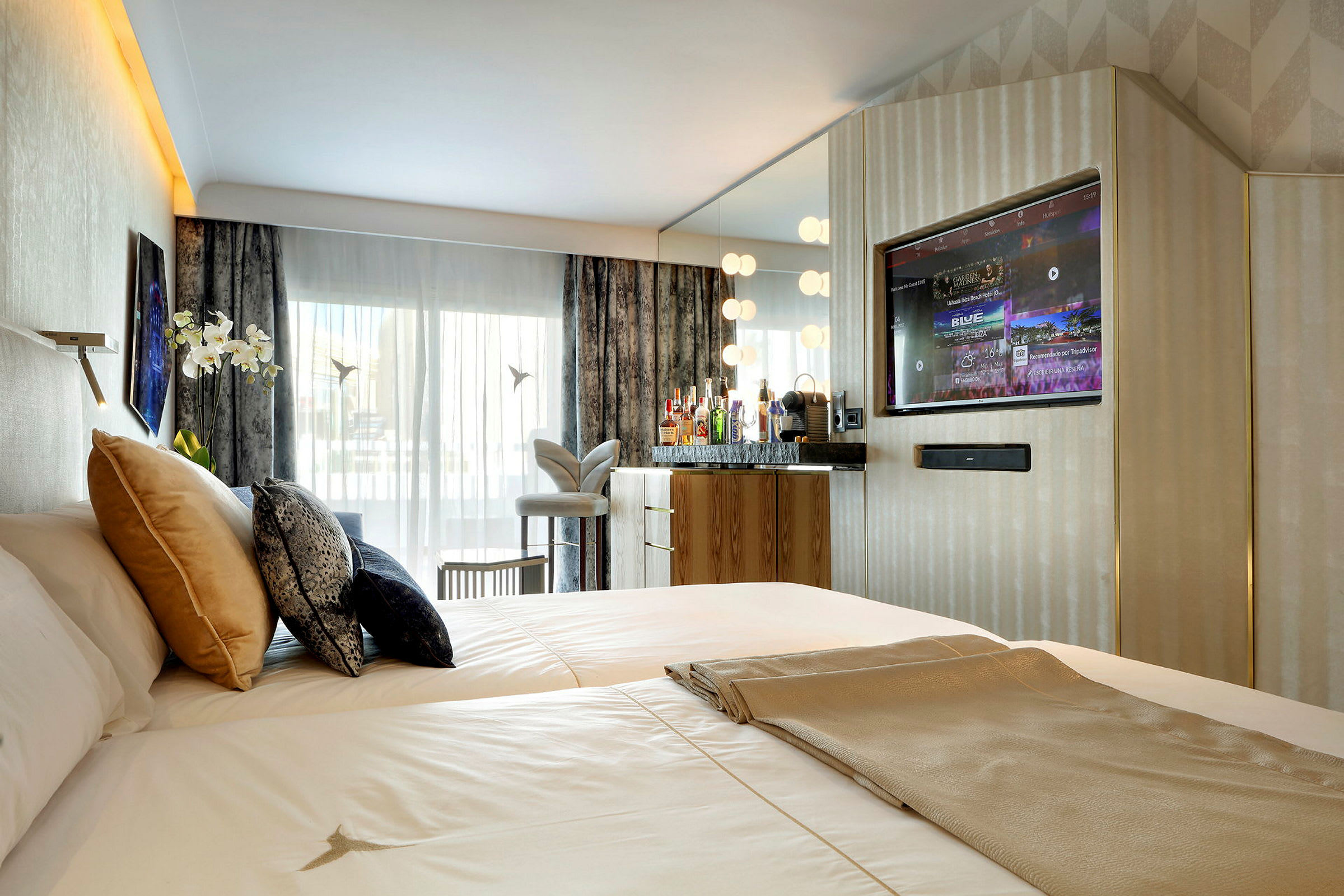 Et hotellrom med en bred seng, flere puter, en TV innebygd i veggen og en speilet benk med drinker og lamper. Store vinduer med gardiner og en dekorativ stol er også synlige. (AI-generert tekst)