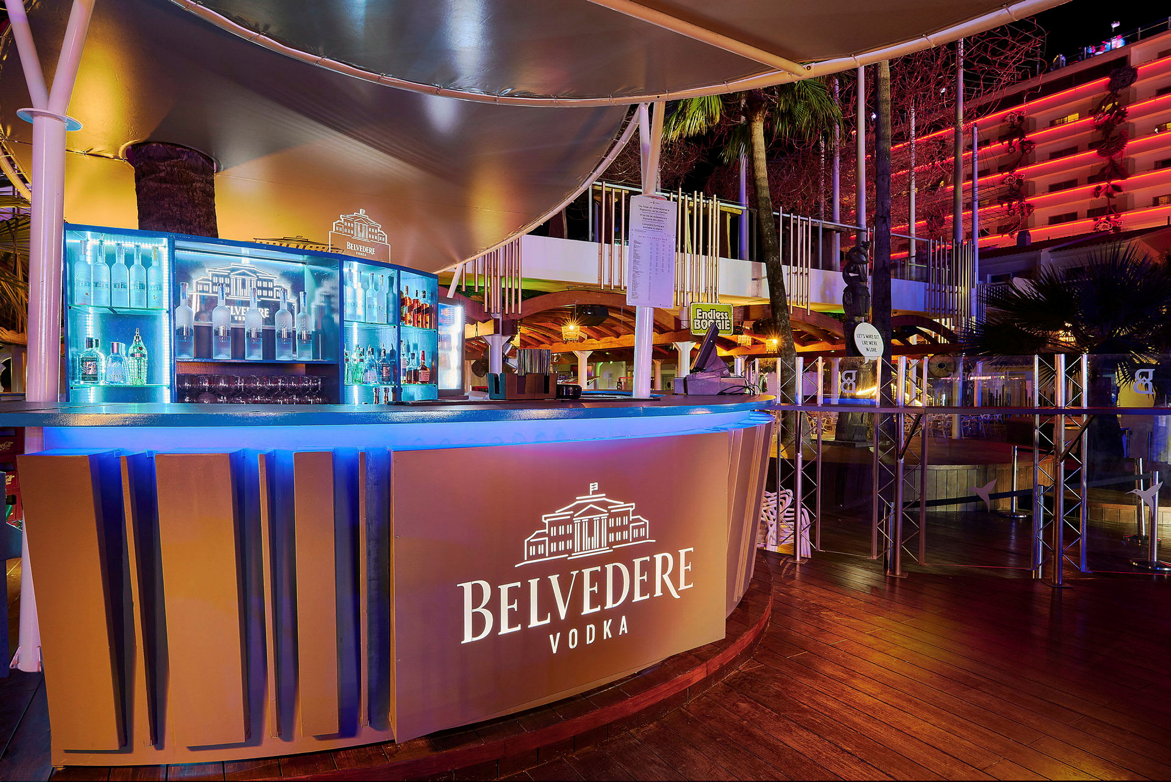 En utendørs bar med skilt for Belvedere Vodka, med belysning og flasker i bakgrunnen. (AI-generert tekst)