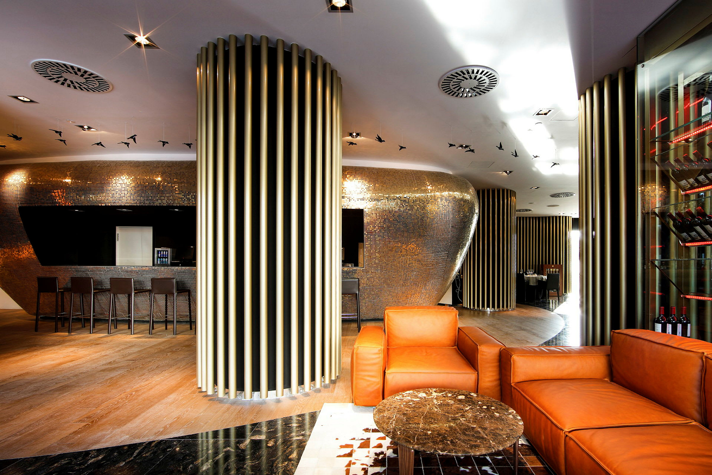 Moderne innredet lounge med oransje lenestoler, et lite bord og metalldekorasjoner på vegger og tak. (AI-generert tekst)