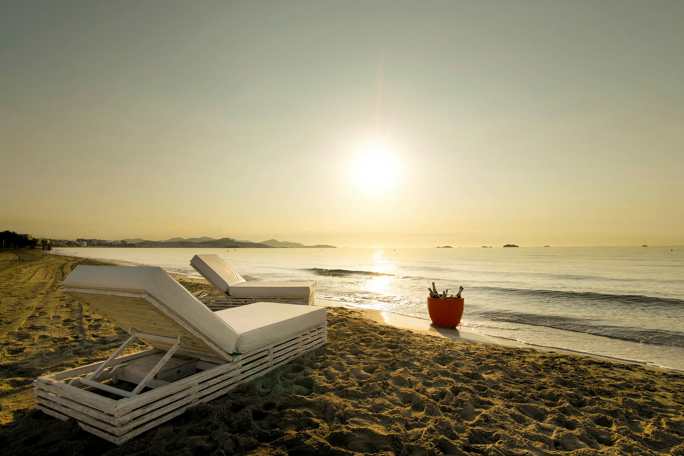 Solstoler på en sandstrand med utsikt over havet ved soloppgang eller solnedgang. En bøtte med flasker står ved siden av. (AI-generert tekst)