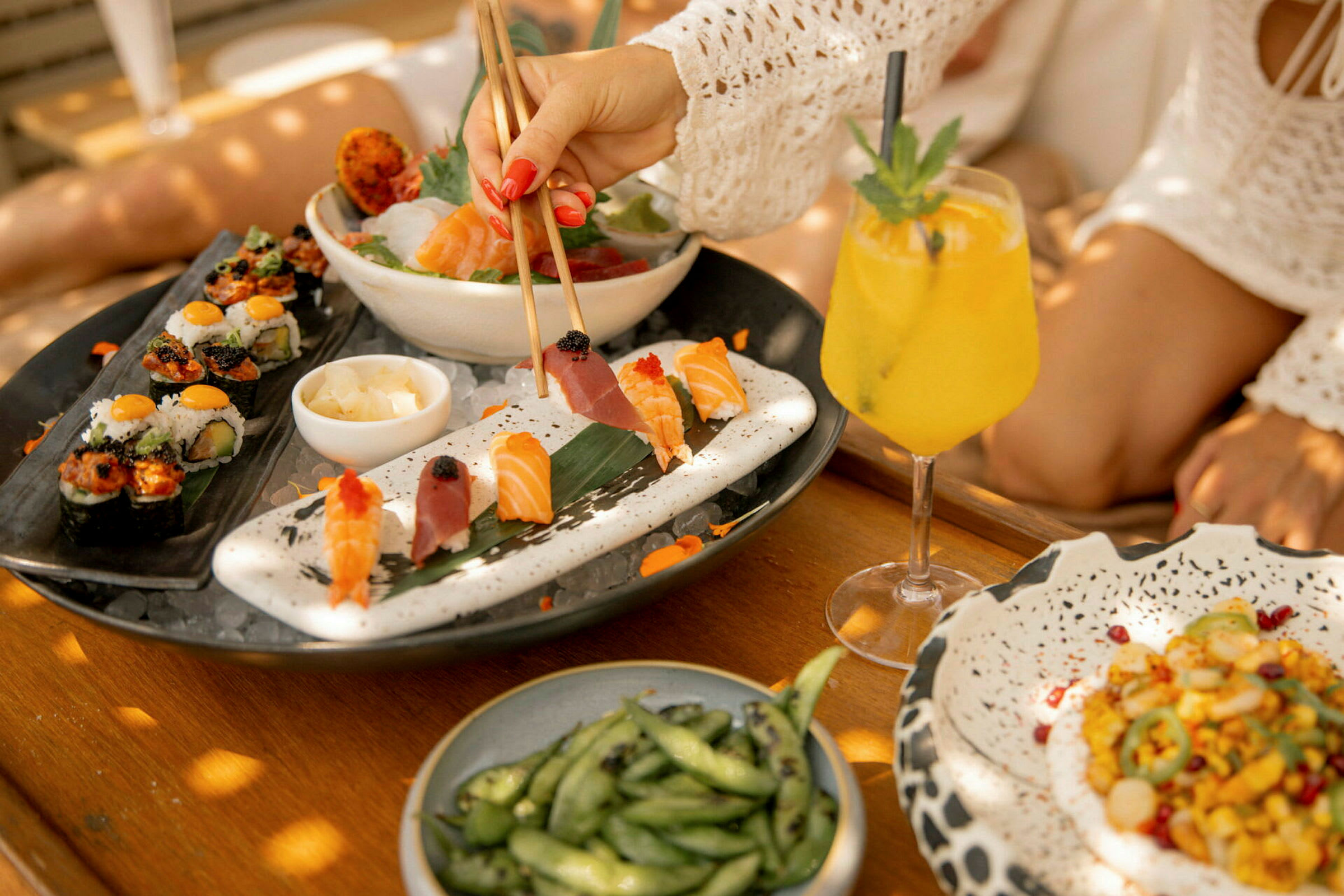 En tallerken med sushi og sashimi sammen med en gul drikk og en bolle med soyabønner som står på et trebord. (AI-generert tekst)