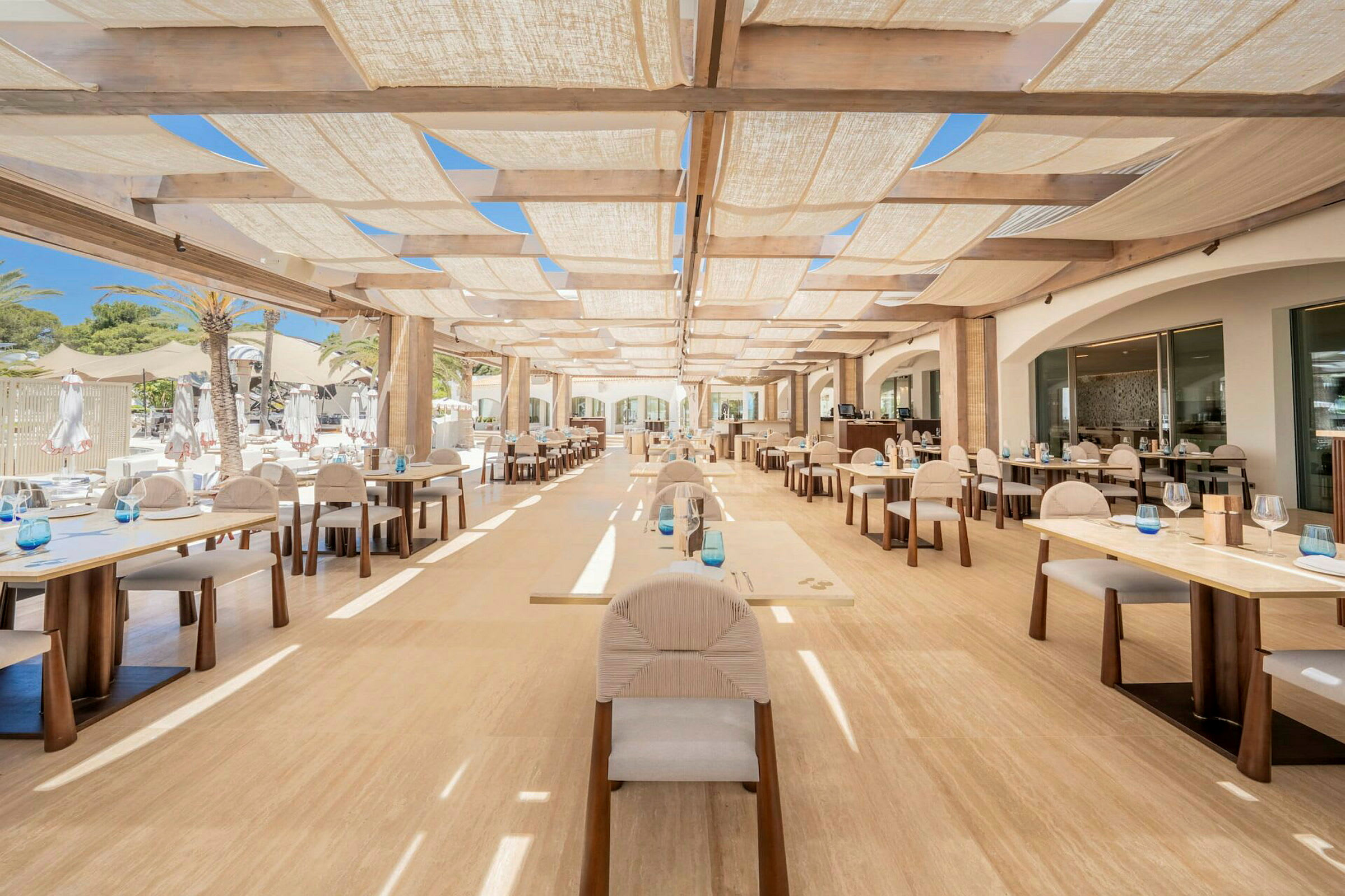 En utendørsrestaurant med bord og stoler under et skyggefullt tak. (AI-generert tekst)