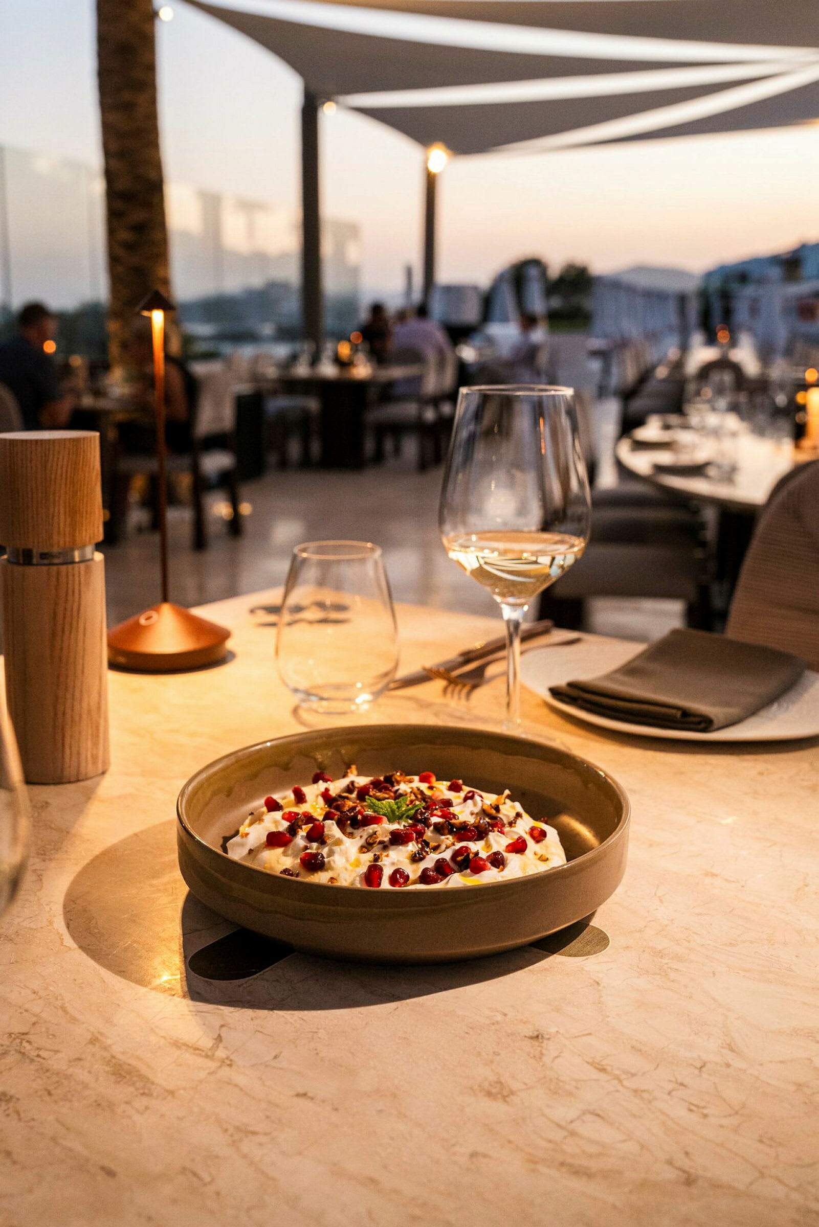 En tallerken med mat, et vinglass og bordtilbehør på et elegant dekket bord, med bakgrunn som viser en restaurantmiljø ved skumring. (AI-generert tekst)