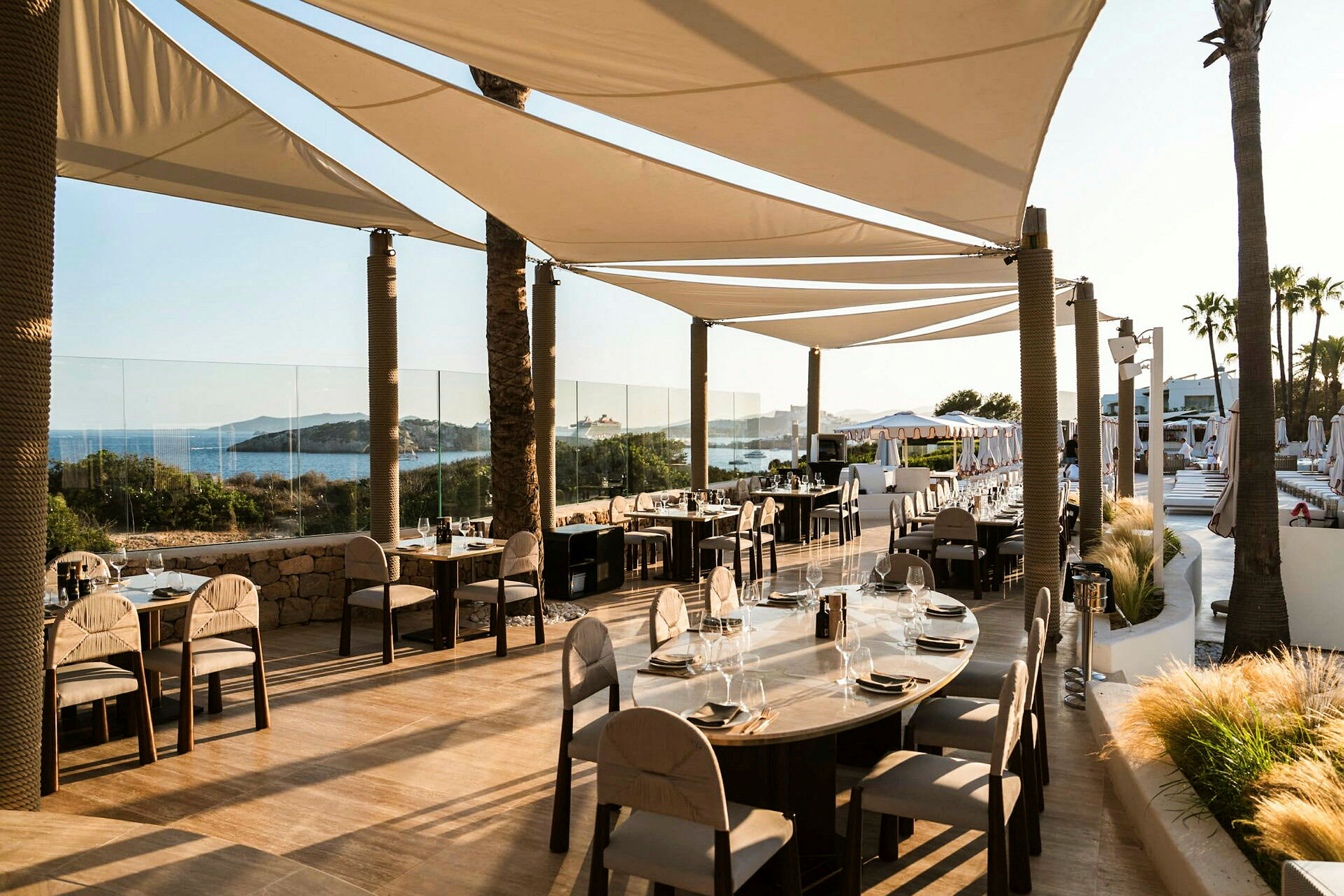 Restaurant med bord og stoler på en terrasse nær havet, skyggelagt av seilduk. (AI-generert tekst)