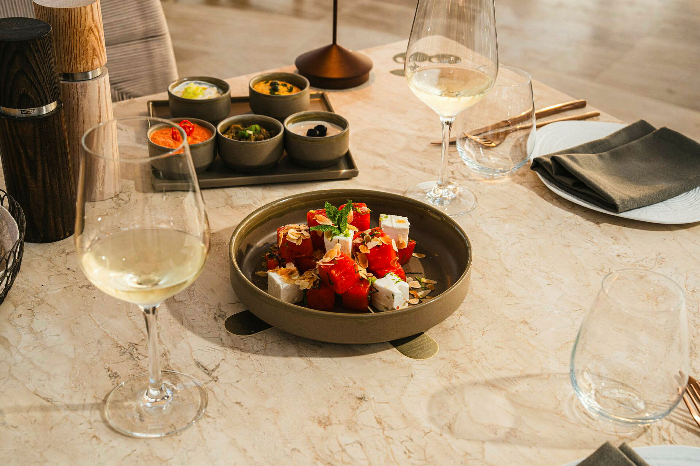 Et bord med en tallerken som inneholder salat med vannmelon og fetaost, omgitt av forskjellige tilbehør i små skåler samt glass med hvitvin. (AI-generert tekst)