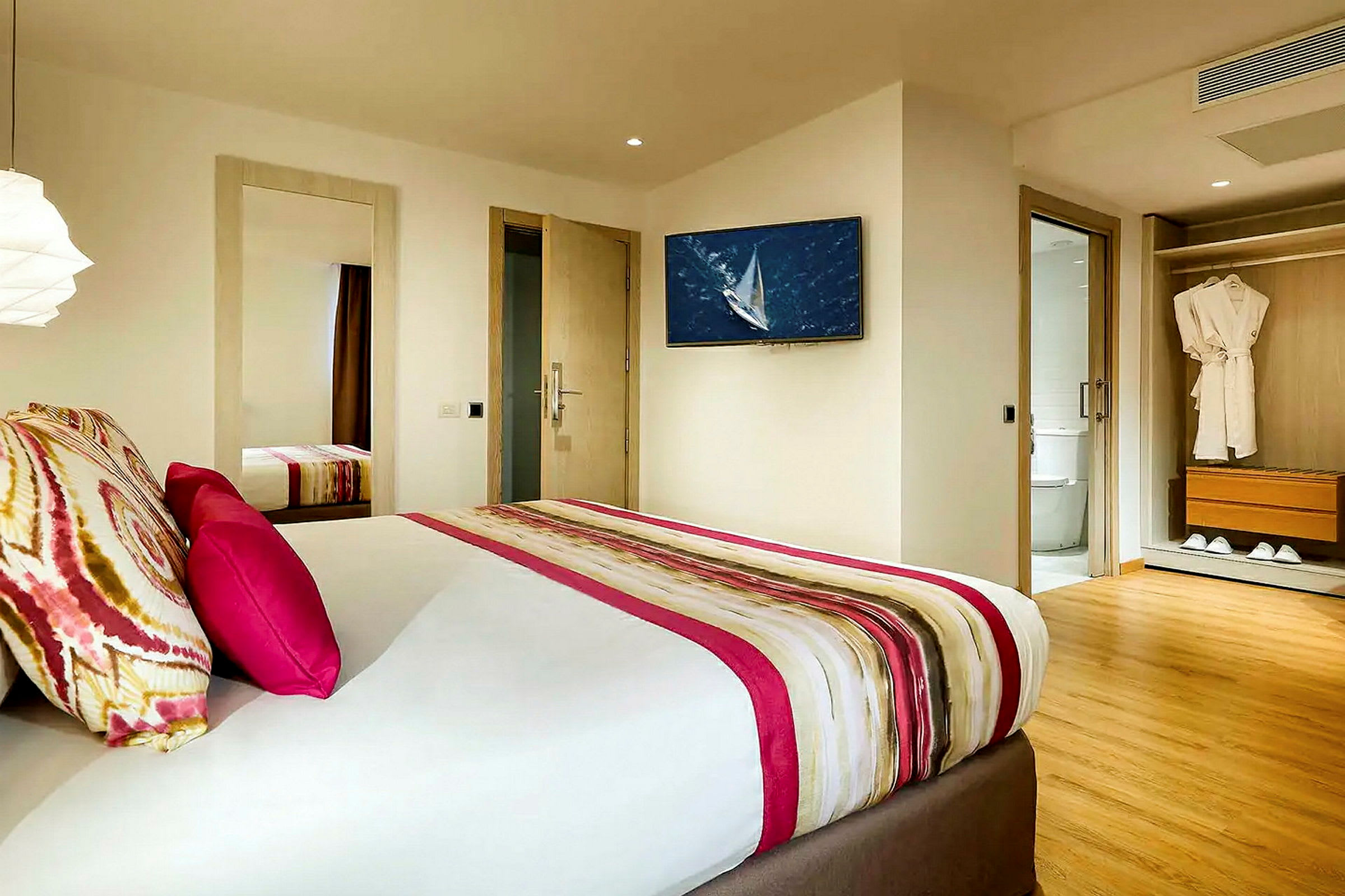Et hotellrom med moderne innredning, en seng med fargerike puter og et teppe, en veggmontert TV, et tilknyttet bad og en garderobe med badekåper. (AI-generert tekst)