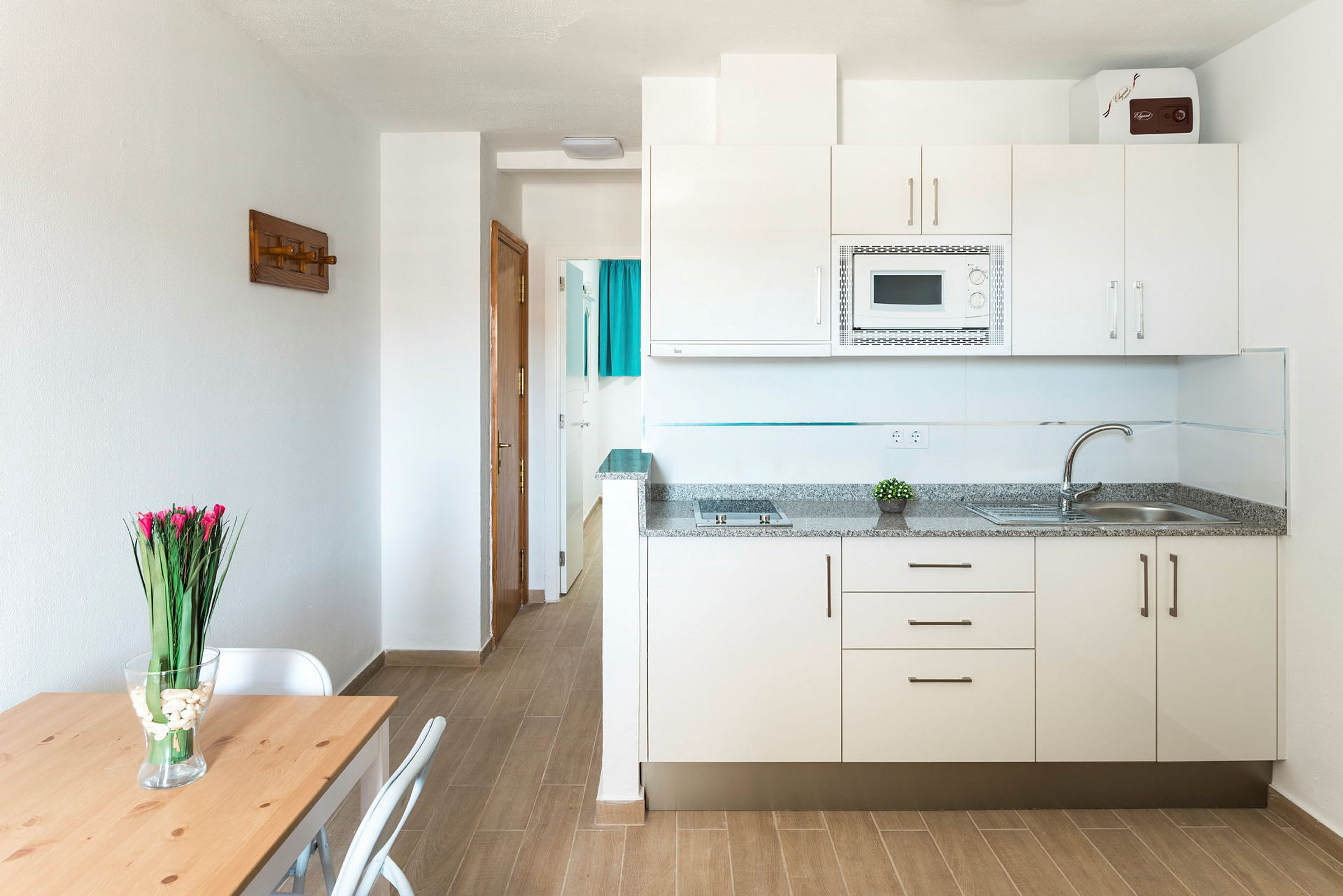 Et moderne kjøkkendel med hvite skap og mikrobølgeovn, ved siden av en liten spisekrok med et bord, to stoler og en vase med blomster. (AI-generert tekst)