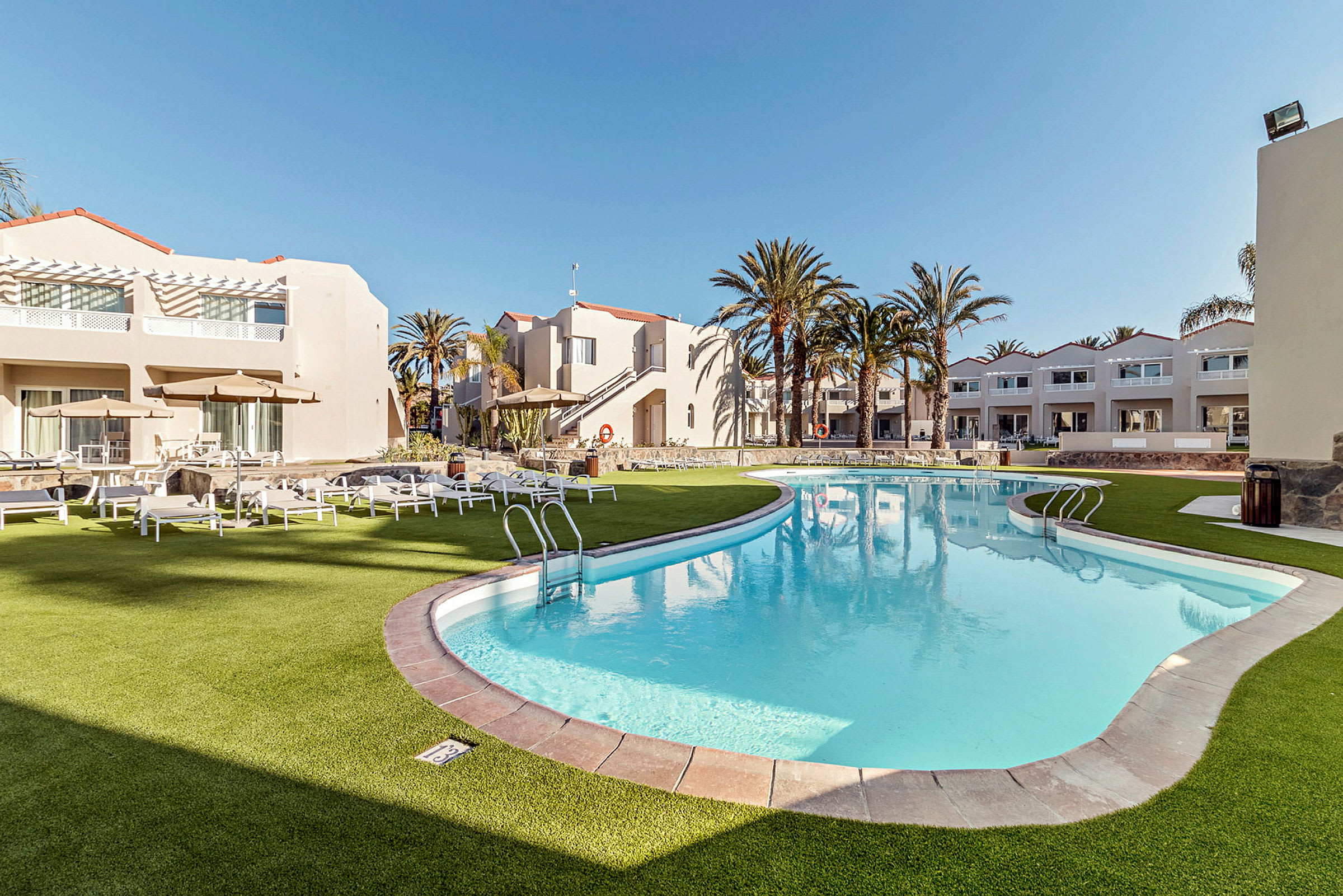 Et utendørs basseng omgitt av hvite bygninger og solsenger på gress. (AI-generert tekst)
