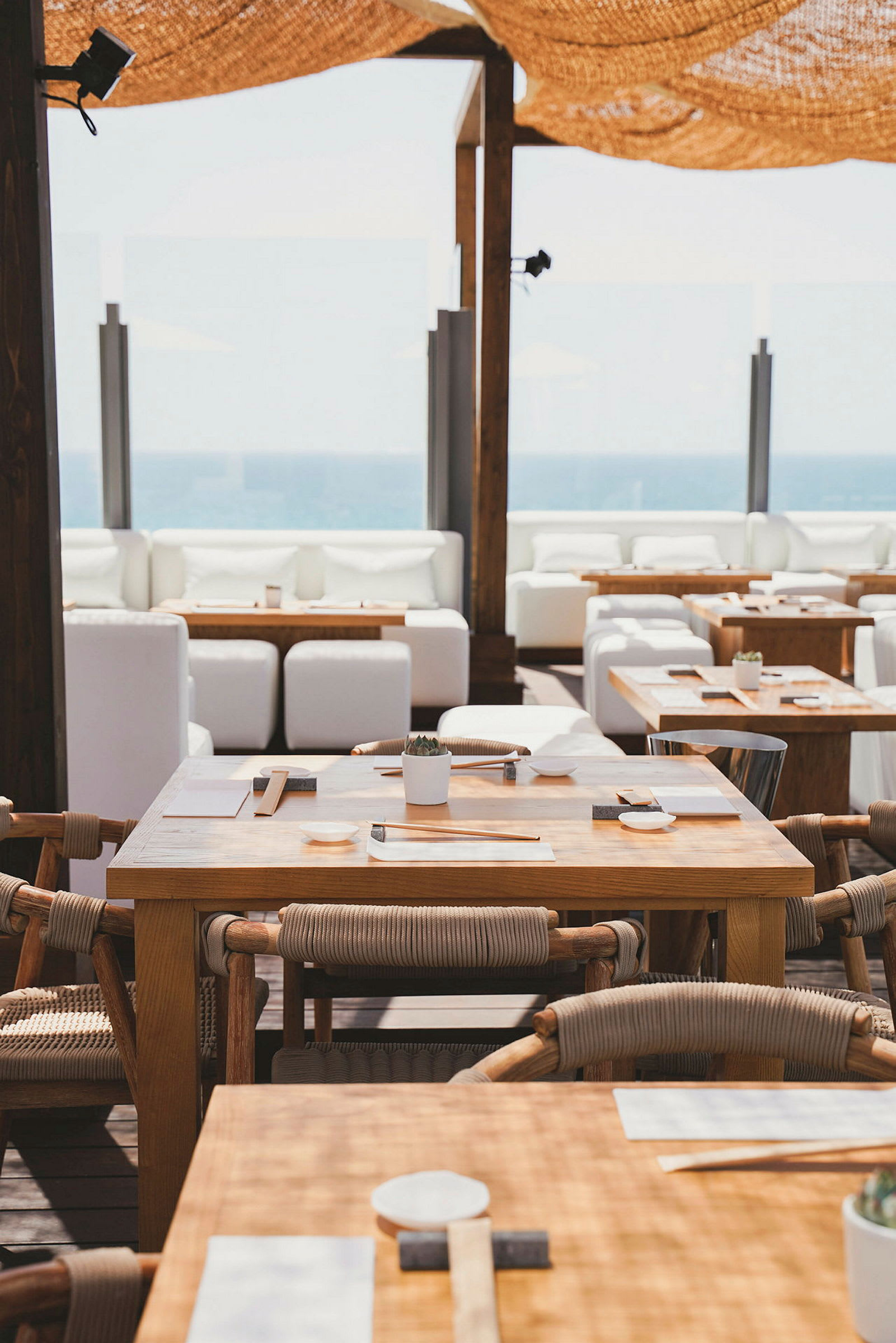 Uterestaurant med trebord og stoler, hvite sofaer i bakgrunnen, samt utsikt mot havet. (AI-generert tekst)