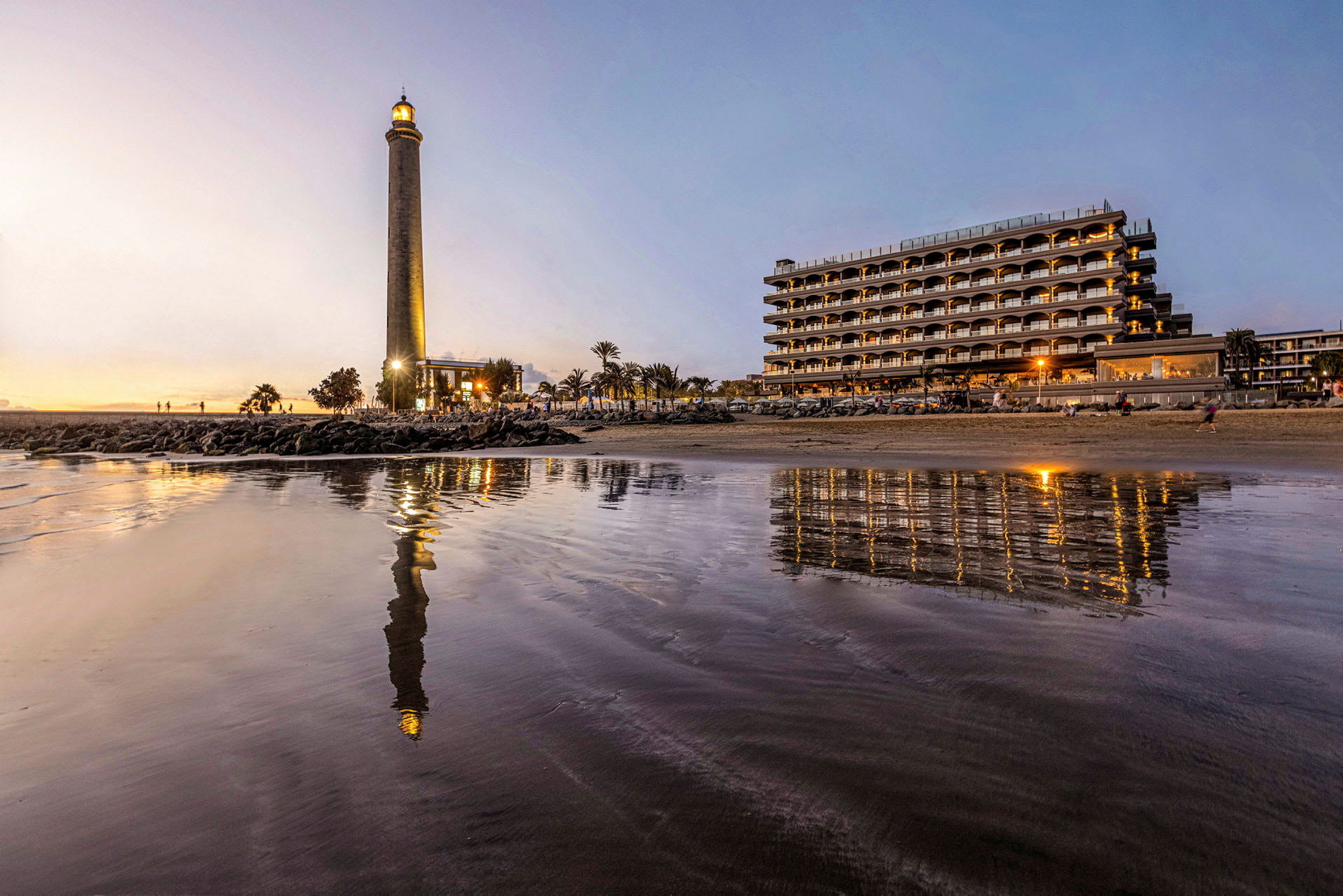 Bilde av et fyrtårn ved stranden med et hotell i bakgrunnen, refleksjoner synlige i vannflaten. (AI-generert tekst)