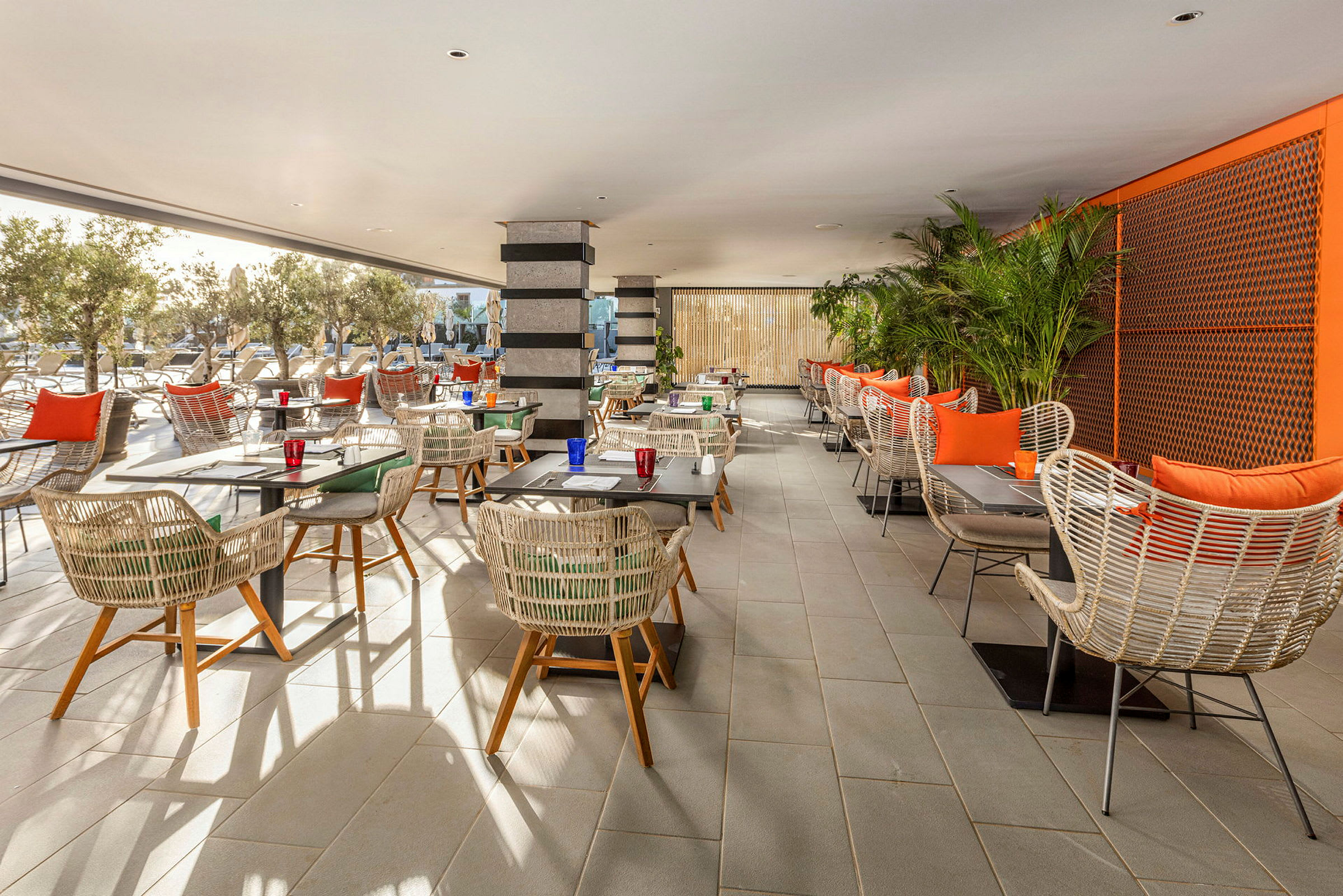 En restaurant med bord og stoler i rottingstil, omgitt av planter og utsikt mot et utendørsområde. (AI-generert tekst)