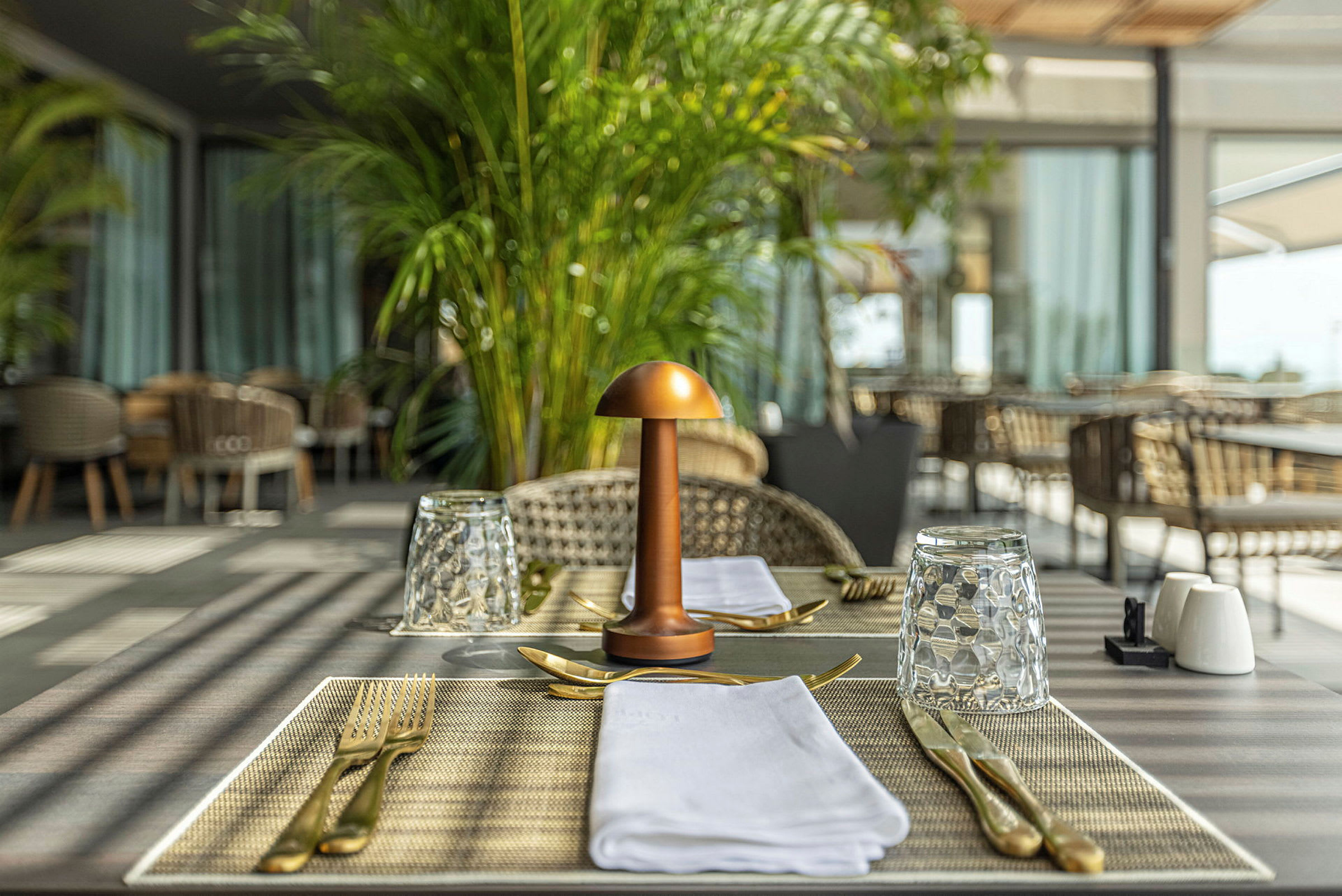 Et bord i et restaurantområde er dekket med glass, bestikk og servietter. En dekorativ lampe er plassert i midten. (AI-generert tekst)