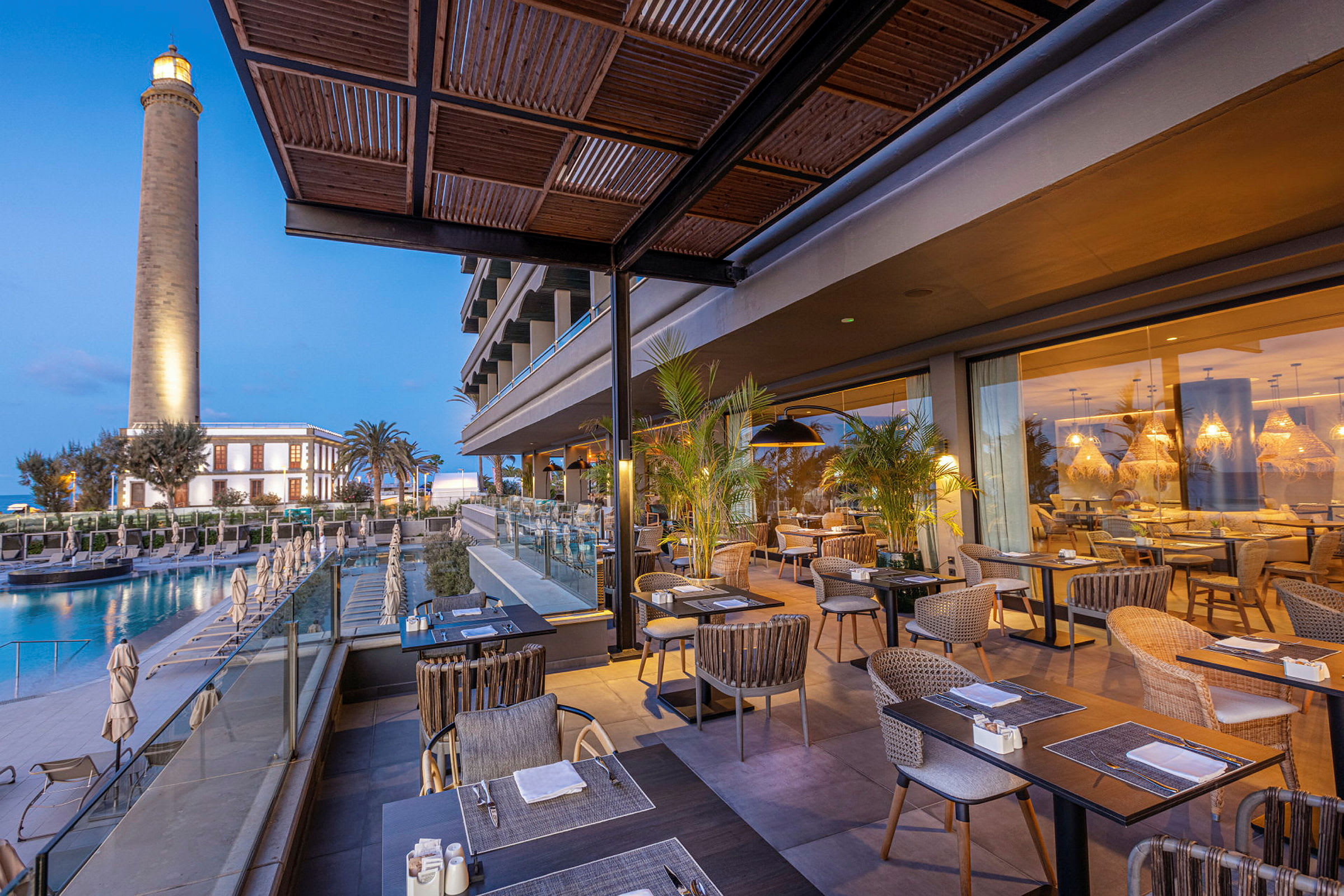 Restaurant med utendørsbord ved et hotell, et fyrtårn i bakgrunnen. (AI-generert tekst)