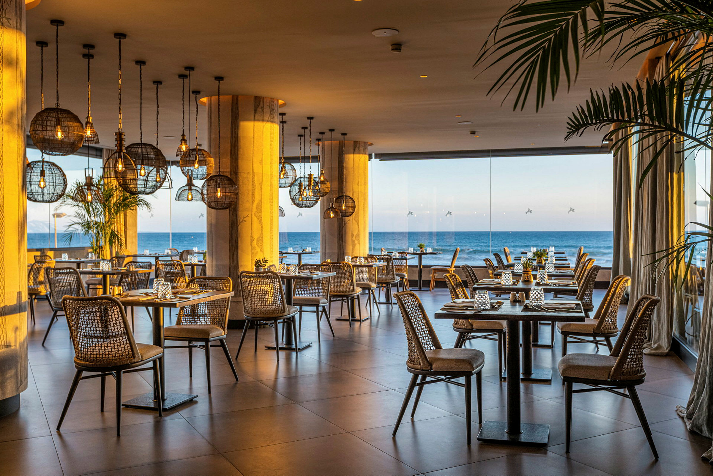 Bilde av en restaurant med rottingstoler, bordene er dekket med utsikt over havet gjennom store glassvinduer. (AI-generert tekst)
