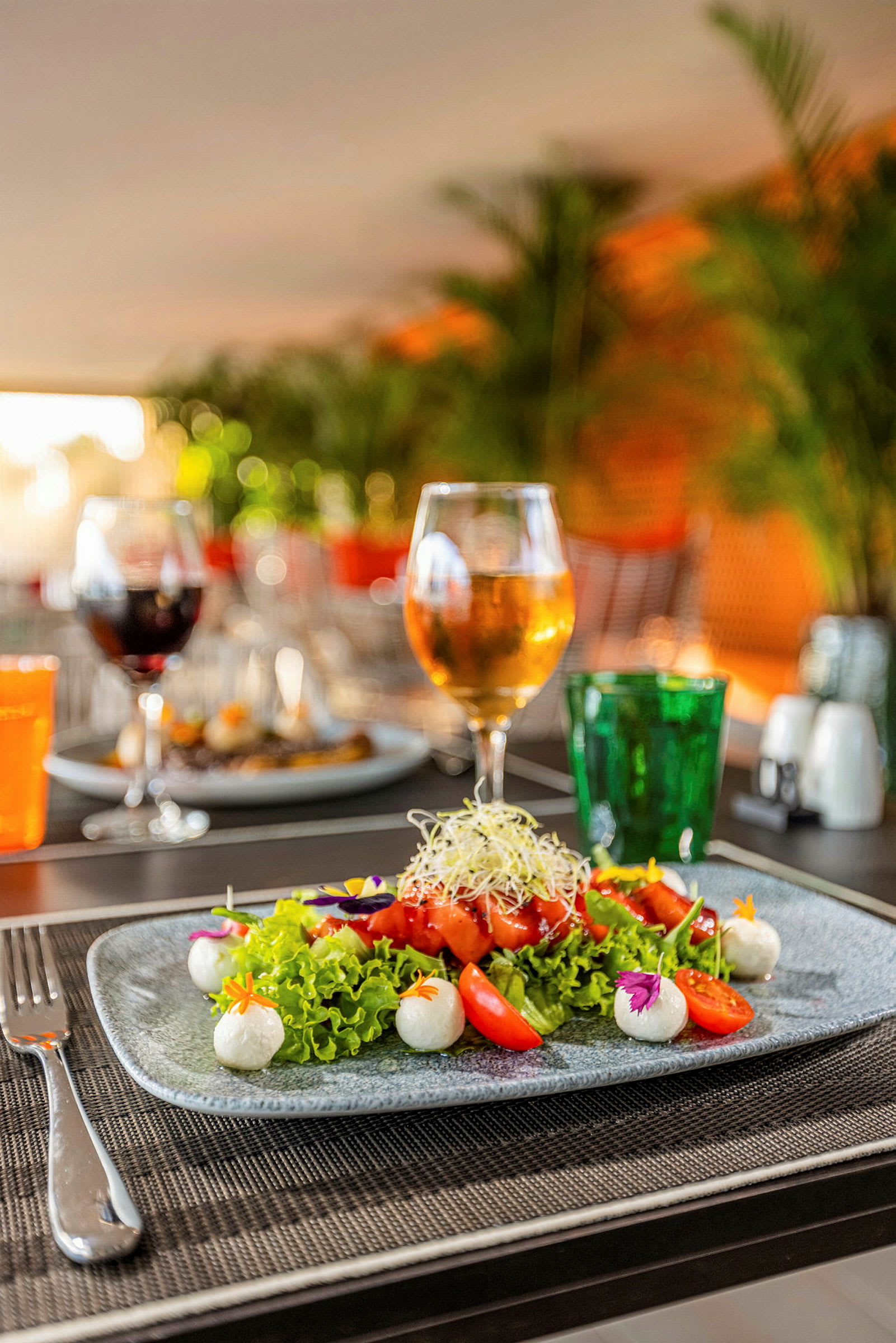 En tallerken med fargerik salat, tomater og mozzarella-kuler står på et dekket bord. Bakgrunnen viser et glass med drikke og grønne planter. (AI-generert tekst)