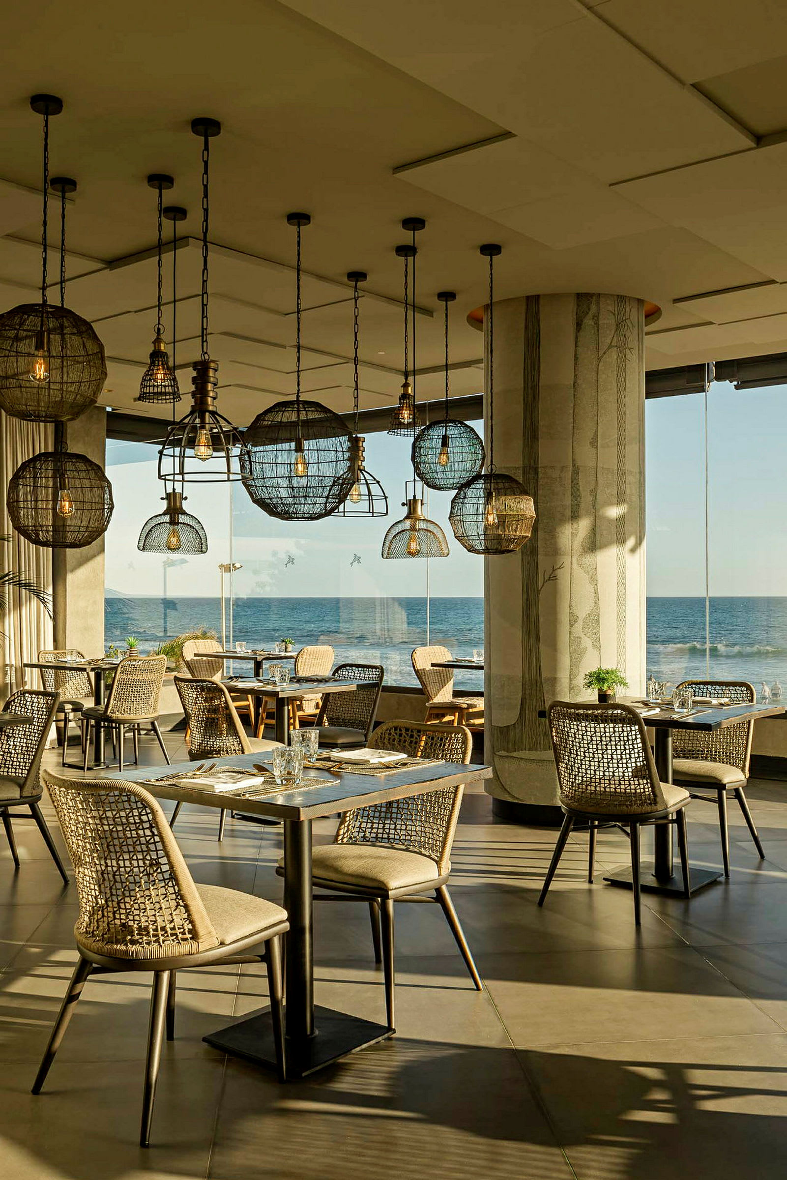 En restaurant med moderne innredning, flere spisebord og stoler samt dekorative lamper i taket. Vinduer med utsikt over havet. (AI-generert tekst)