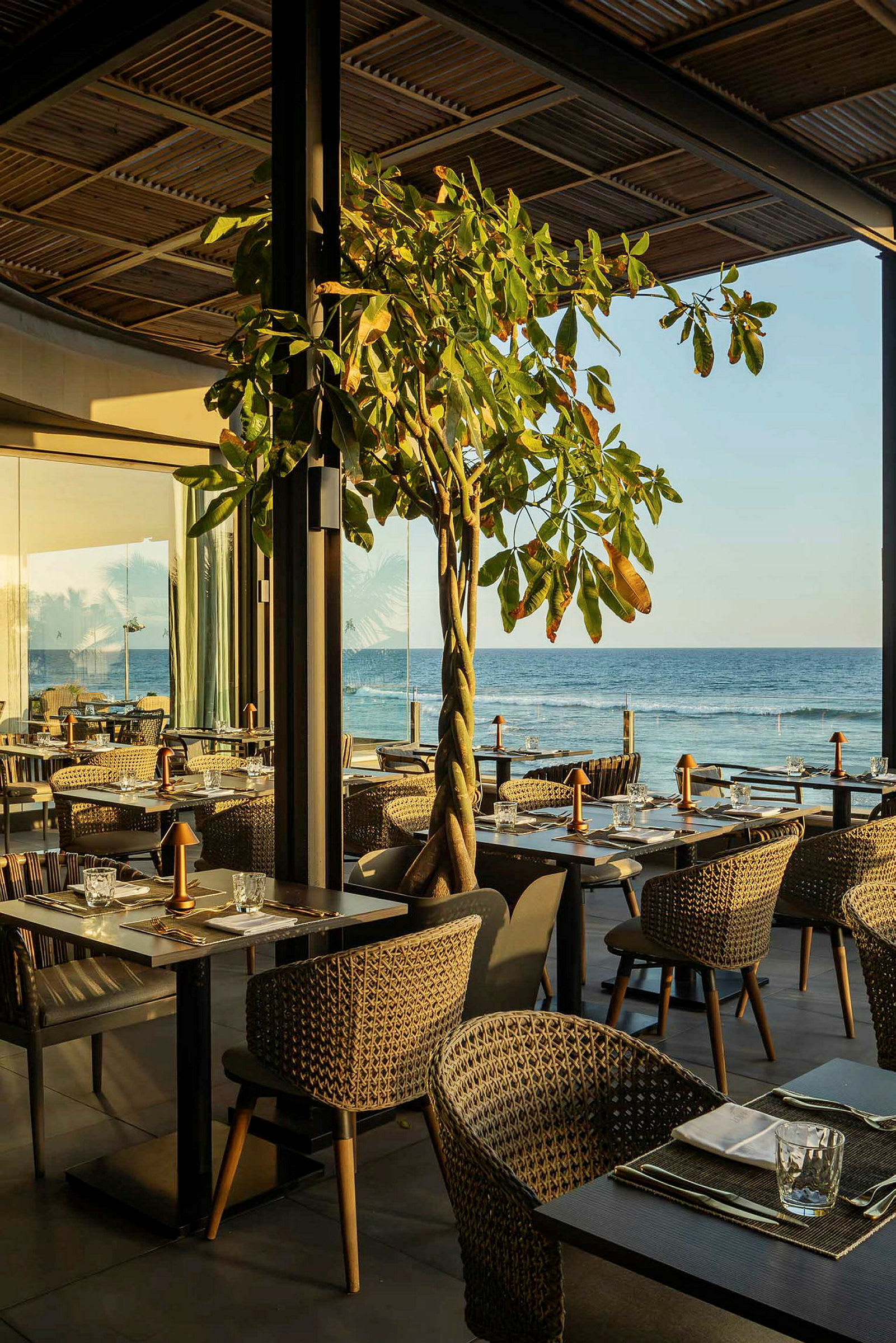 En restaurant med utsikt over havet, med bord og stoler plassert i en åpen miljø med naturlige elementer som et tre. (AI-generert tekst)