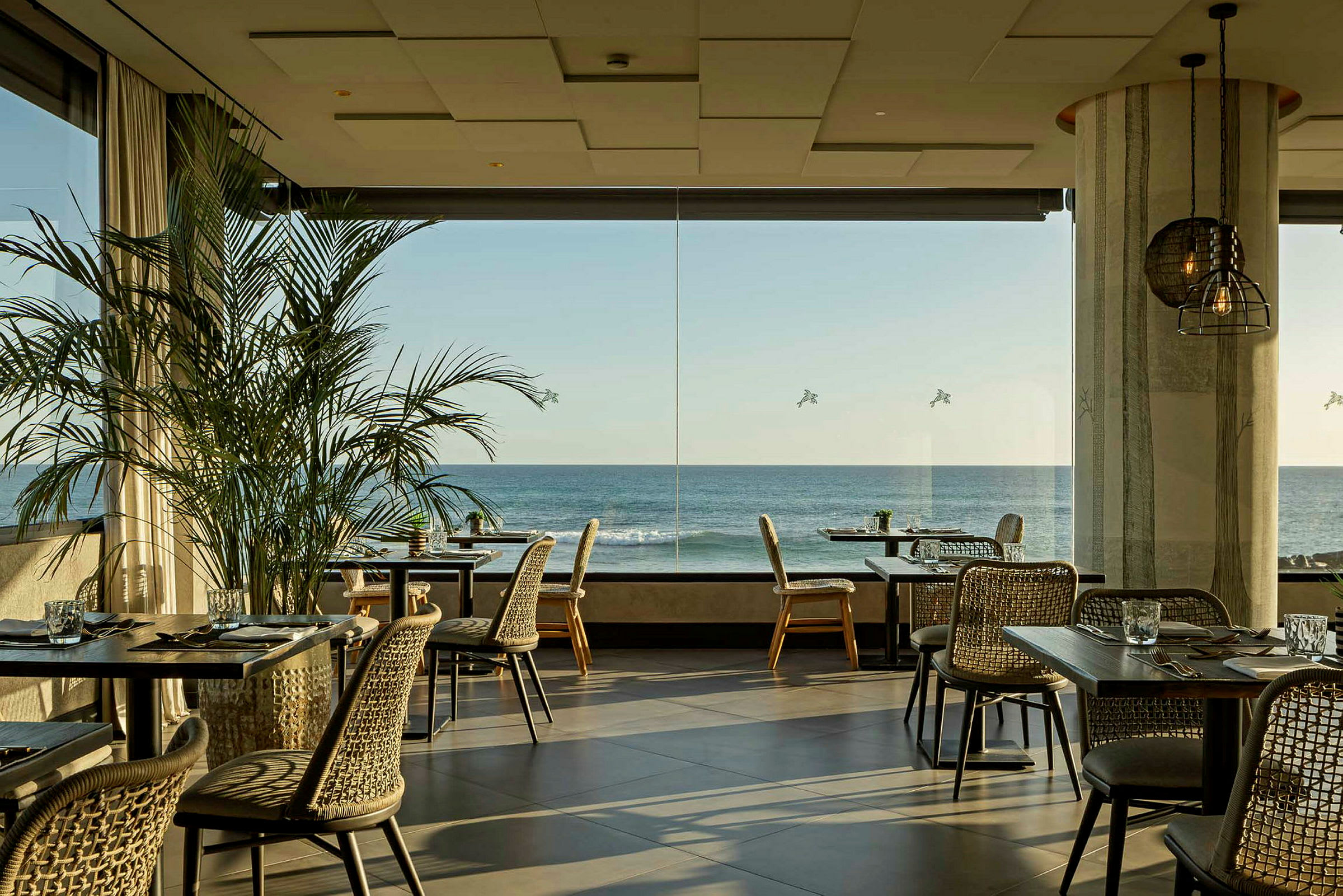 En lys restaurant med store vinduer med utsikt mot havet. Bord og stoler er pent arrangert, og grønne planter pryder interiøret. (AI-generert tekst)