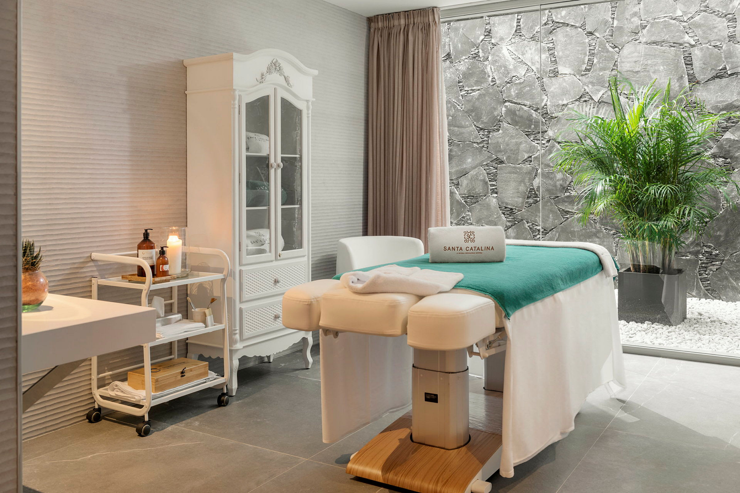 Et elegant spa-rom med massasjebenk, hvit innredning og et stort vindu som vender mot en steinmur og grønne planter. (AI-generert tekst)