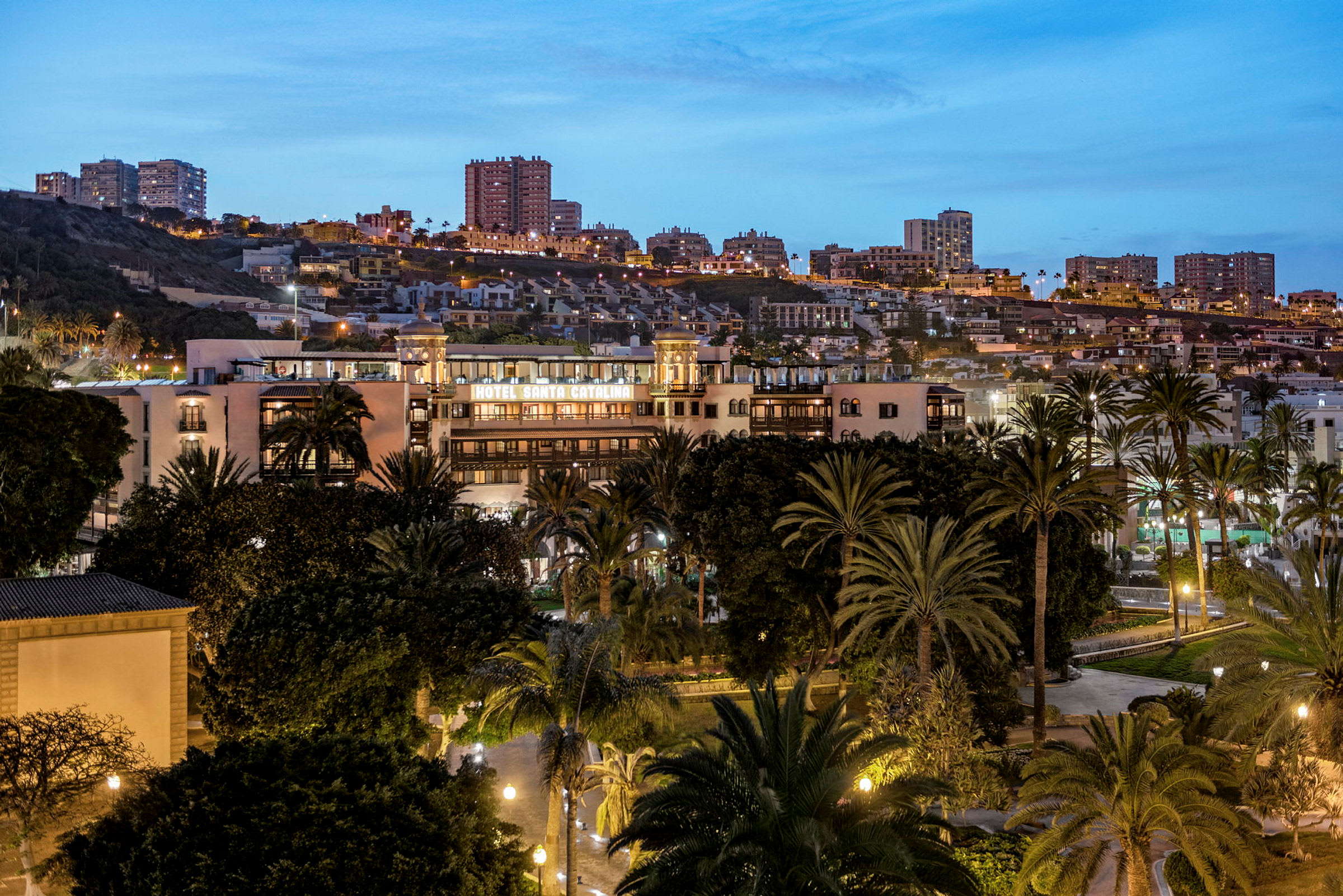 Hotel Santa Catalina omgitt av trær med en bakgrunn av byen om kvelden. (AI-generert tekst)