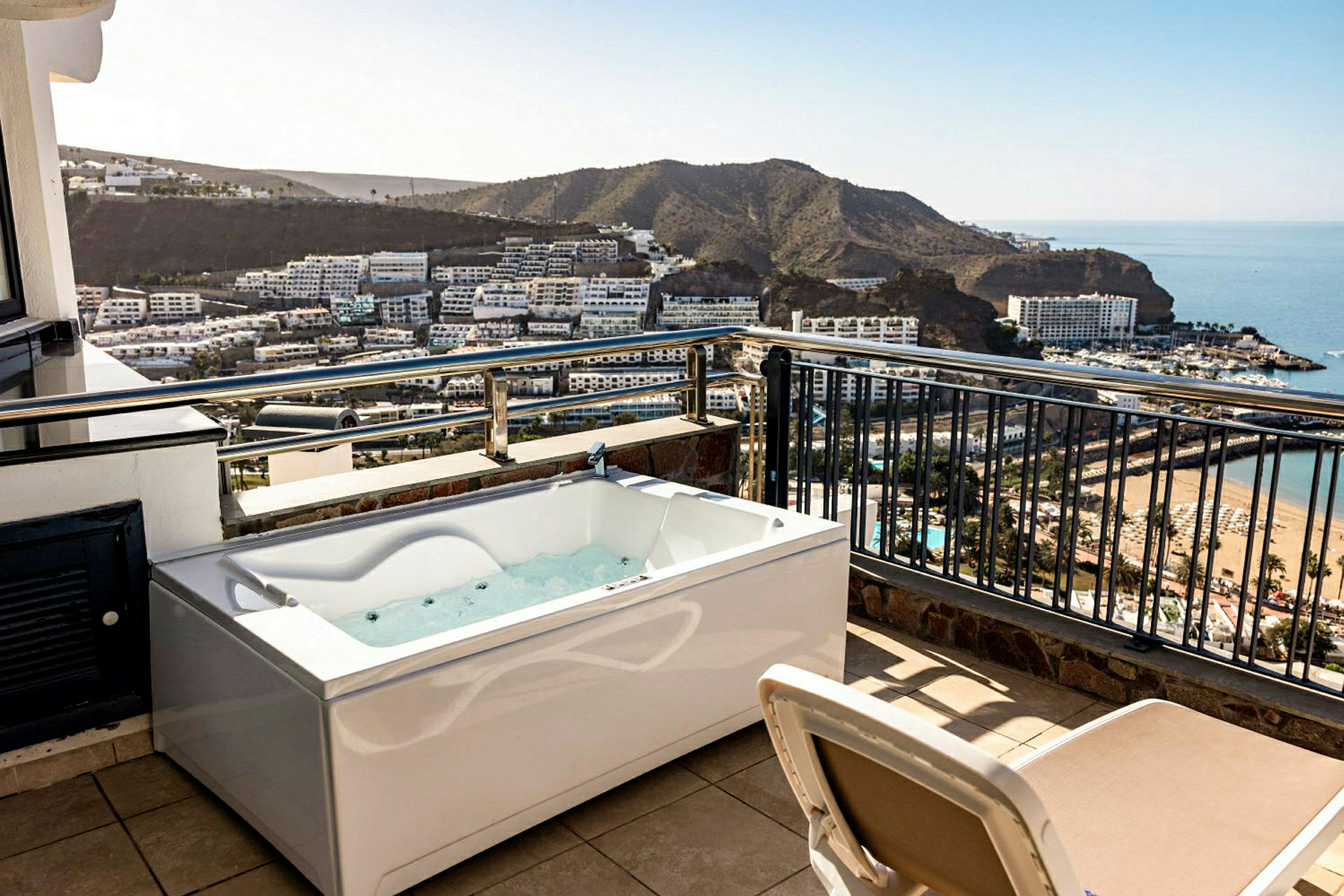En terrasse med boblebad og utsikt over en bukt med hotell og strand. (AI-generert tekst)