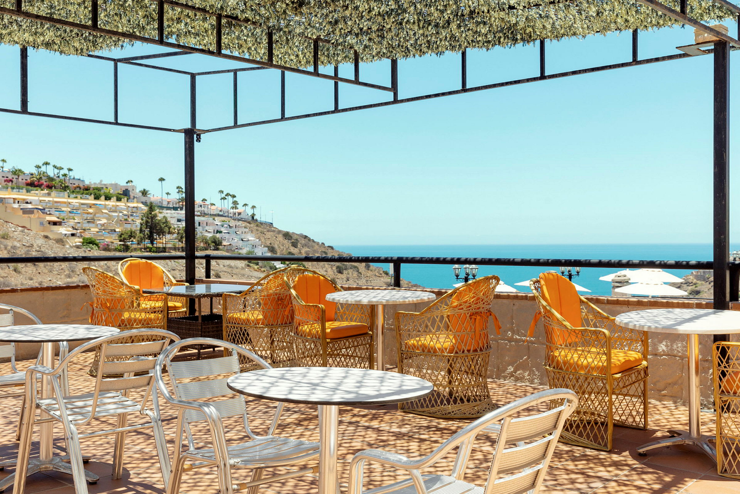 Utendørs servering med bord og stoler på en terrasse med utsikt over havet og en skråning med bygninger. (AI-generert tekst)