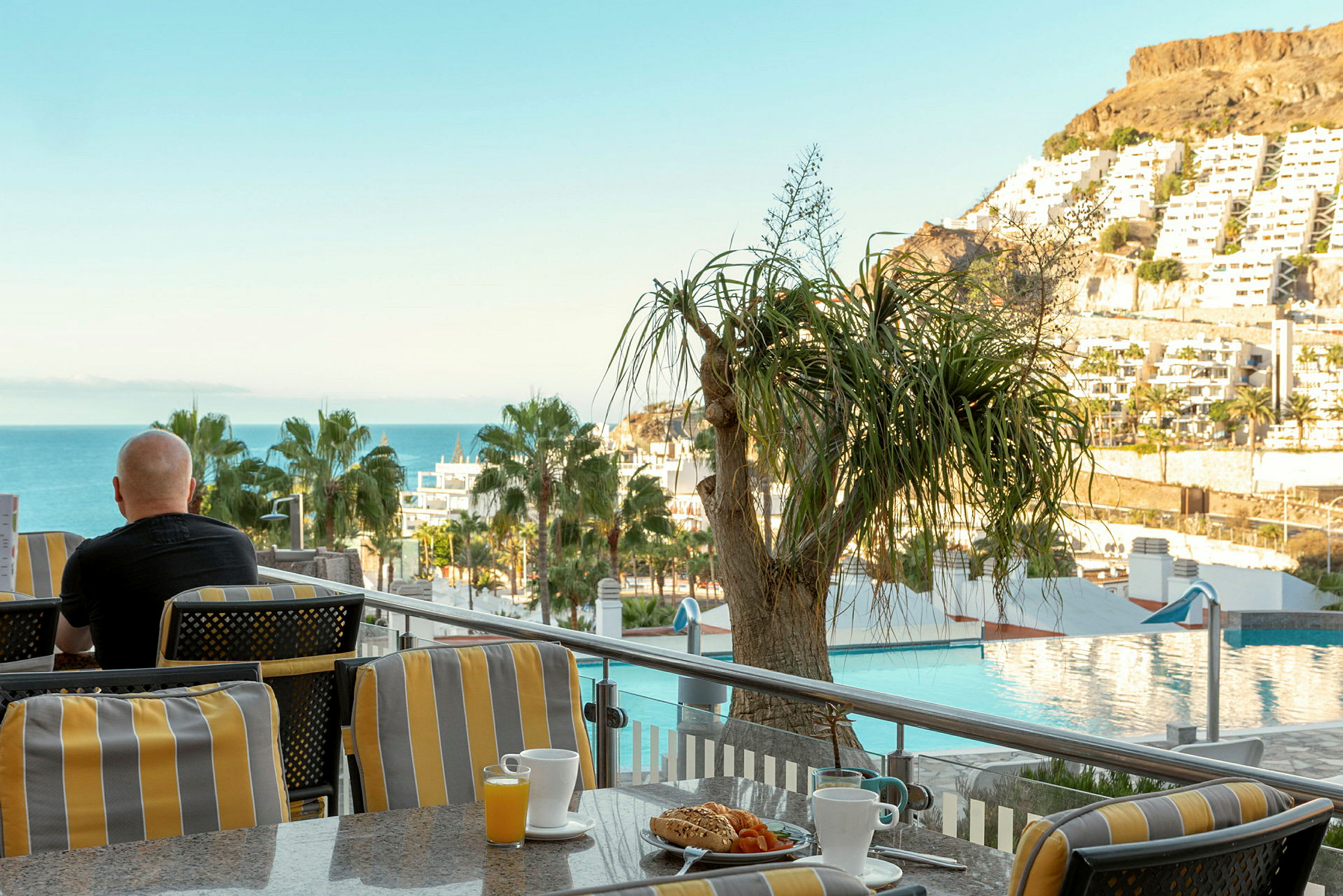 En person sitter på en terrasse med utsikt over havet, trær og bygninger, med en kopp kaffe og frokost foran seg. (AI-generert tekst)