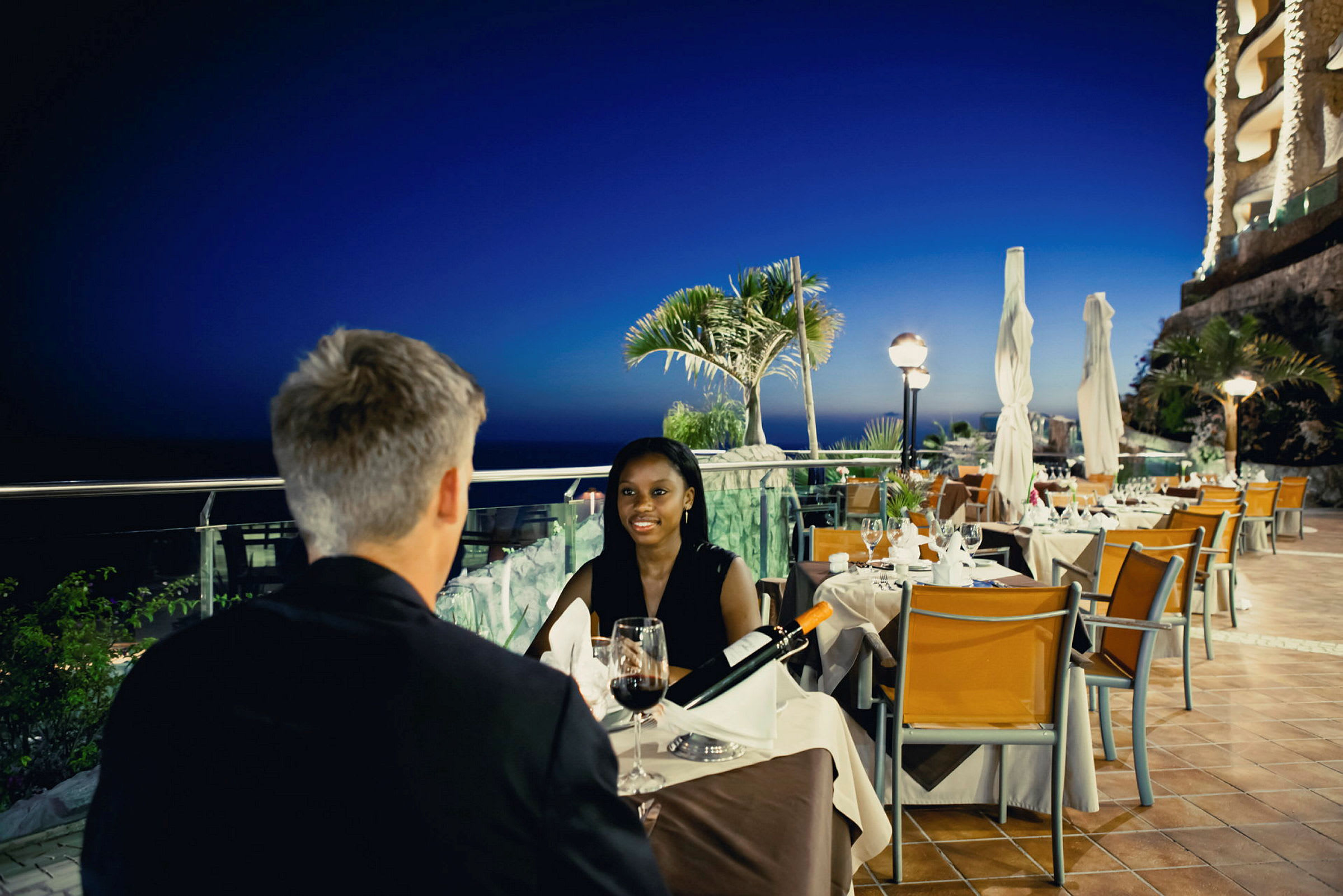 To personer ved et bord på en restaurant med kveldssikt utover havet og tropiske planter. (AI-generert tekst)