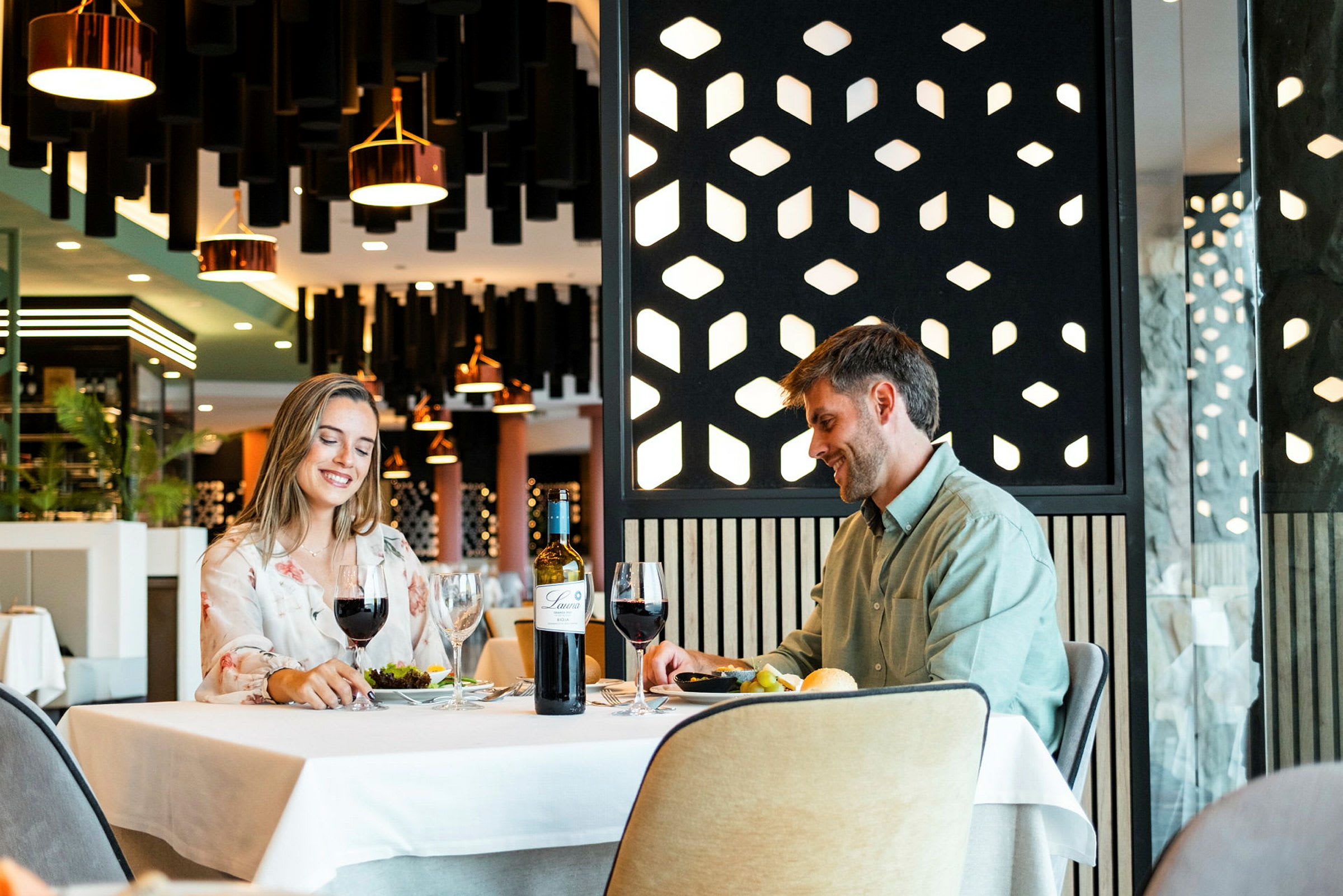 To personer sitter ved et bord på en restaurant med moderne innredning. På bordet er det glass, en vinflaske og tallerkener med mat. (AI-generert tekst)