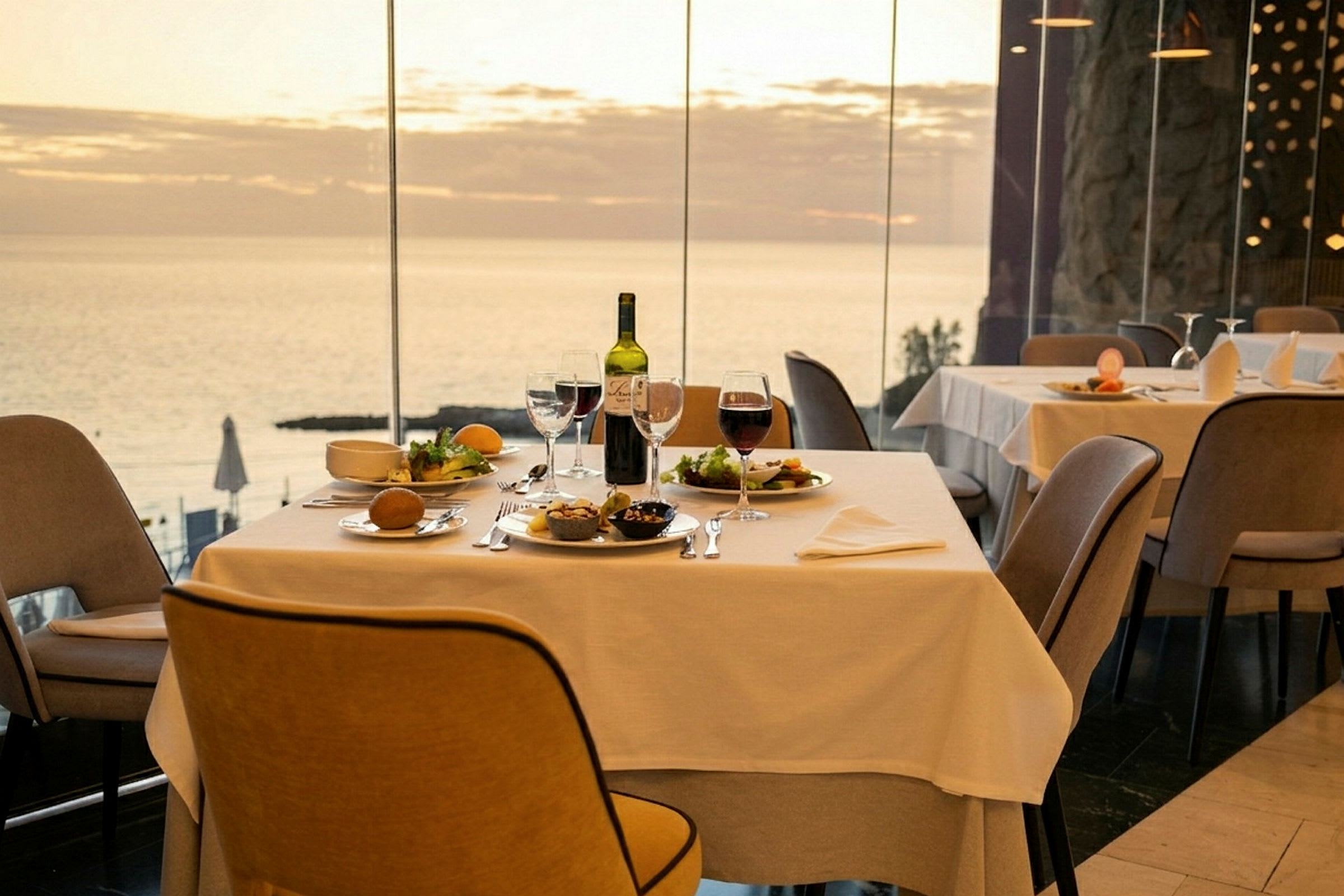 Et bord på en restaurant med hvite duker, servering av mat og vin på bordet, med havutsikt gjennom store vinduer. (AI-generert tekst)